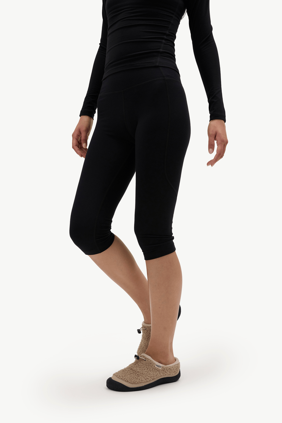 A model wearing black high-waisted capri leggings, a black long-sleeve top, and beige slip-on slippers against a plain white studio background.