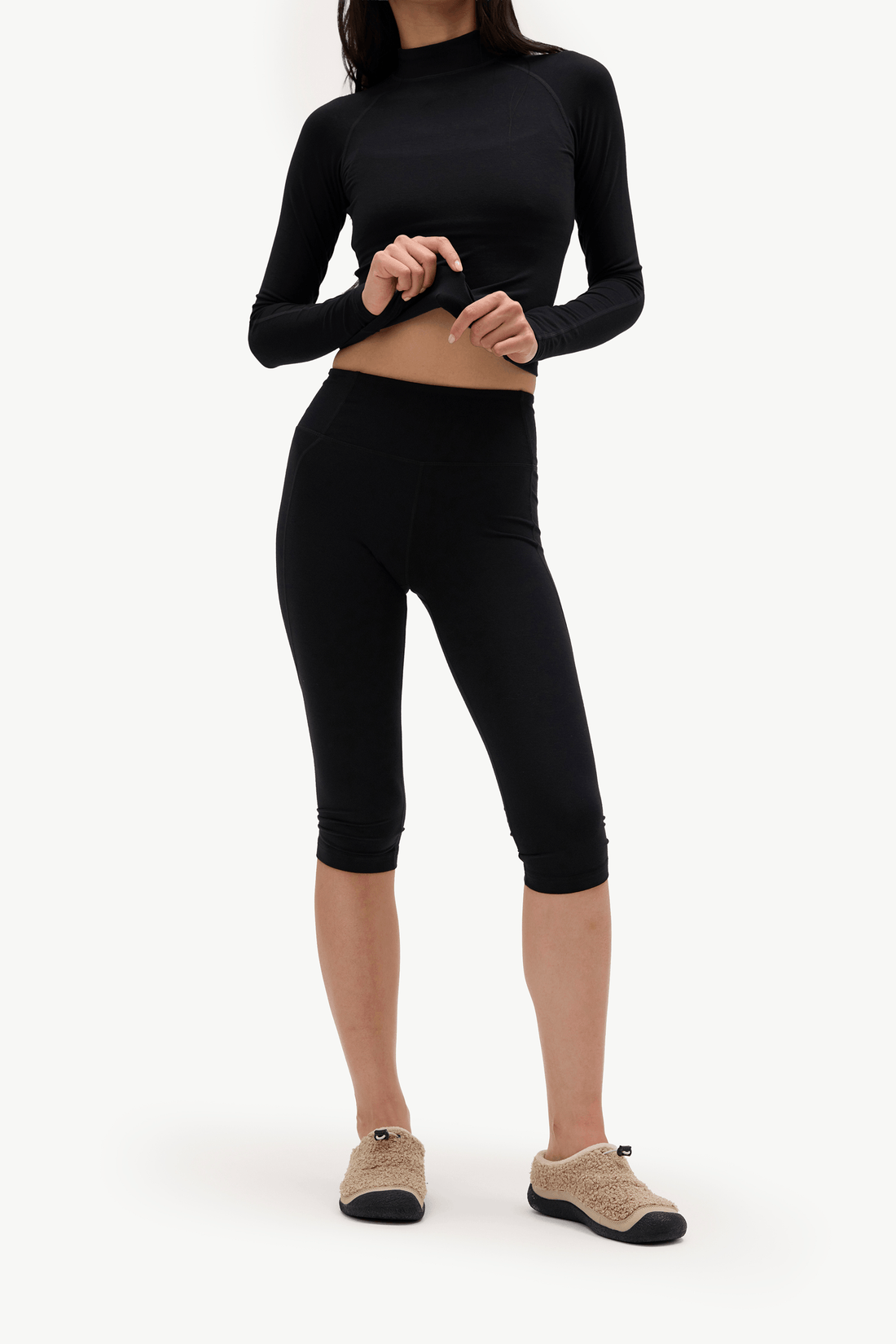 A model wearing a black long-sleeve crop top and matching black capri leggings, paired with beige slip-on shoes, against a plain white studio background.