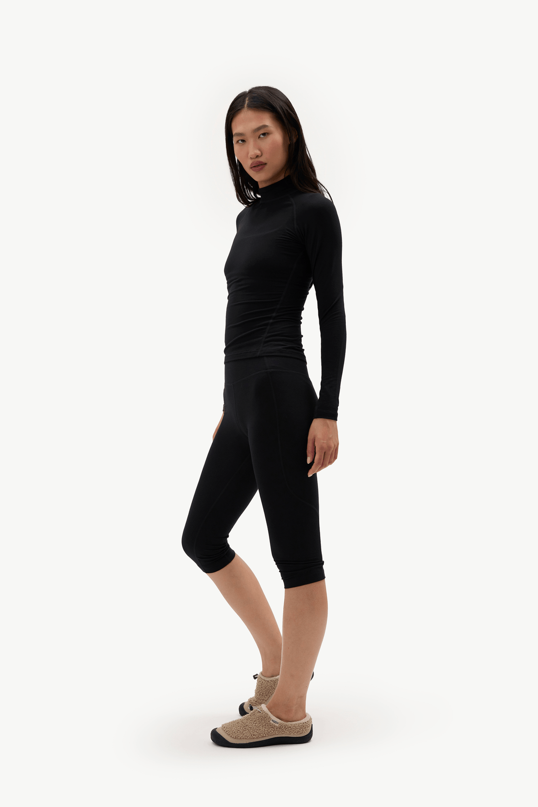 A woman modeling a black long-sleeve mock neck top and matching black capri leggings against a plain white studio background.