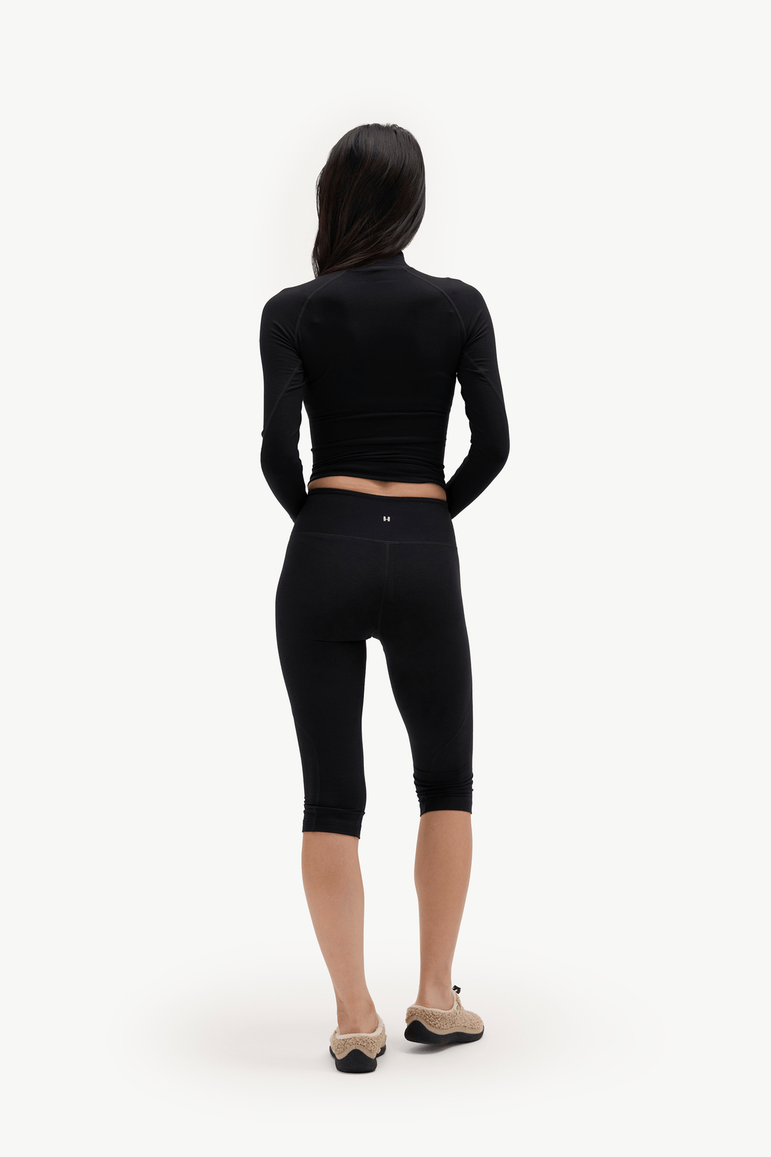A woman viewed from behind wearing a black long-sleeve cropped top and black capri leggings, paired with beige flats, against a plain white studio background.