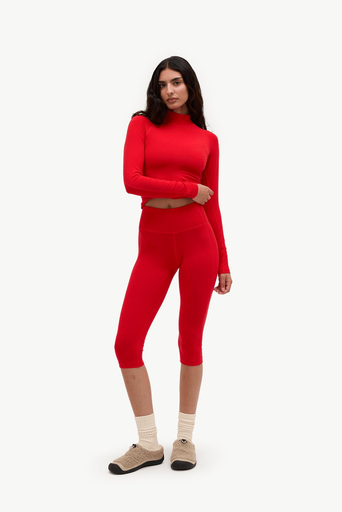 A woman modeling a matching red two-piece activewear set consisting of a long-sleeve crop top and high-waisted capri leggings, paired with beige socks and brown slip-on shoes, against a plain white background.