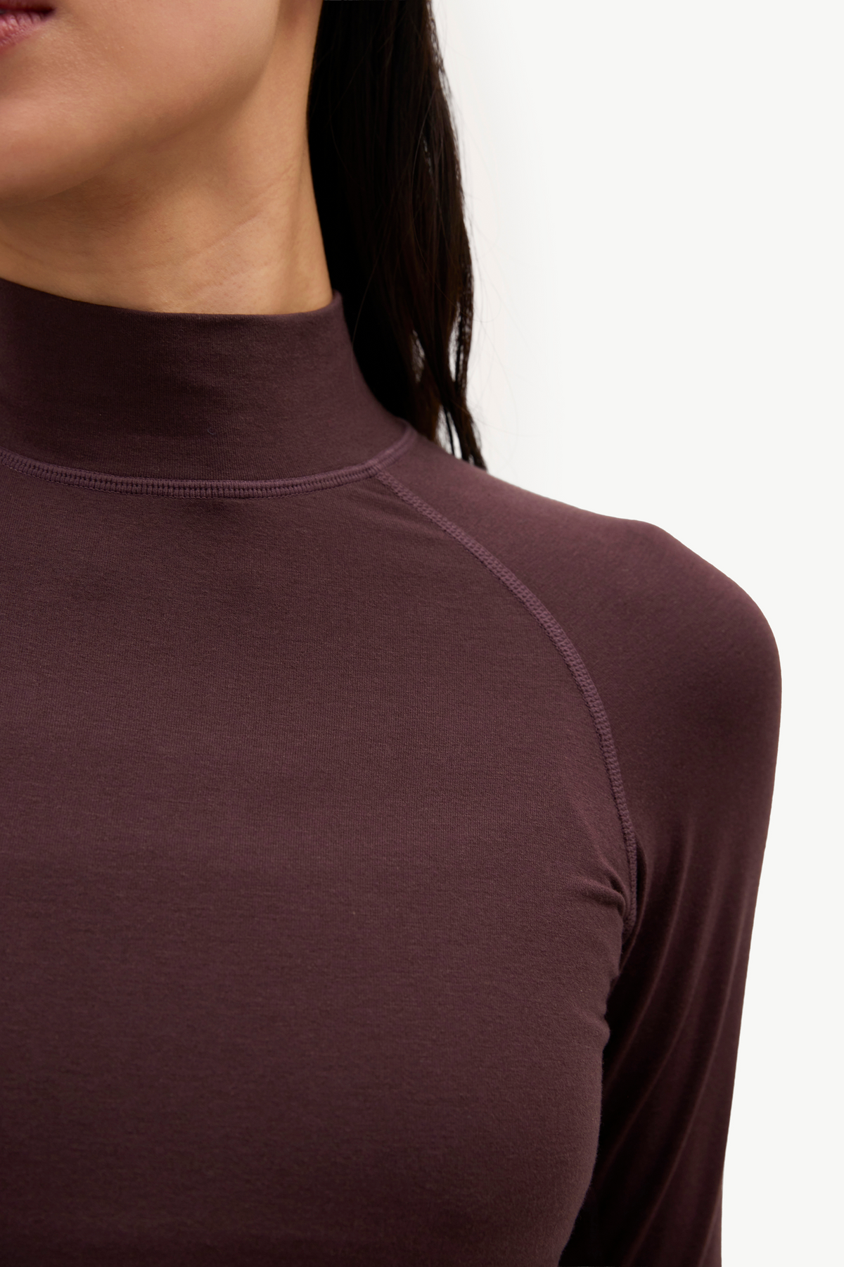 Close-up of a person wearing a fitted long-sleeve mock neck top in a dark plum-brown color, showing the neck, shoulder, and upper chest against a plain light background.