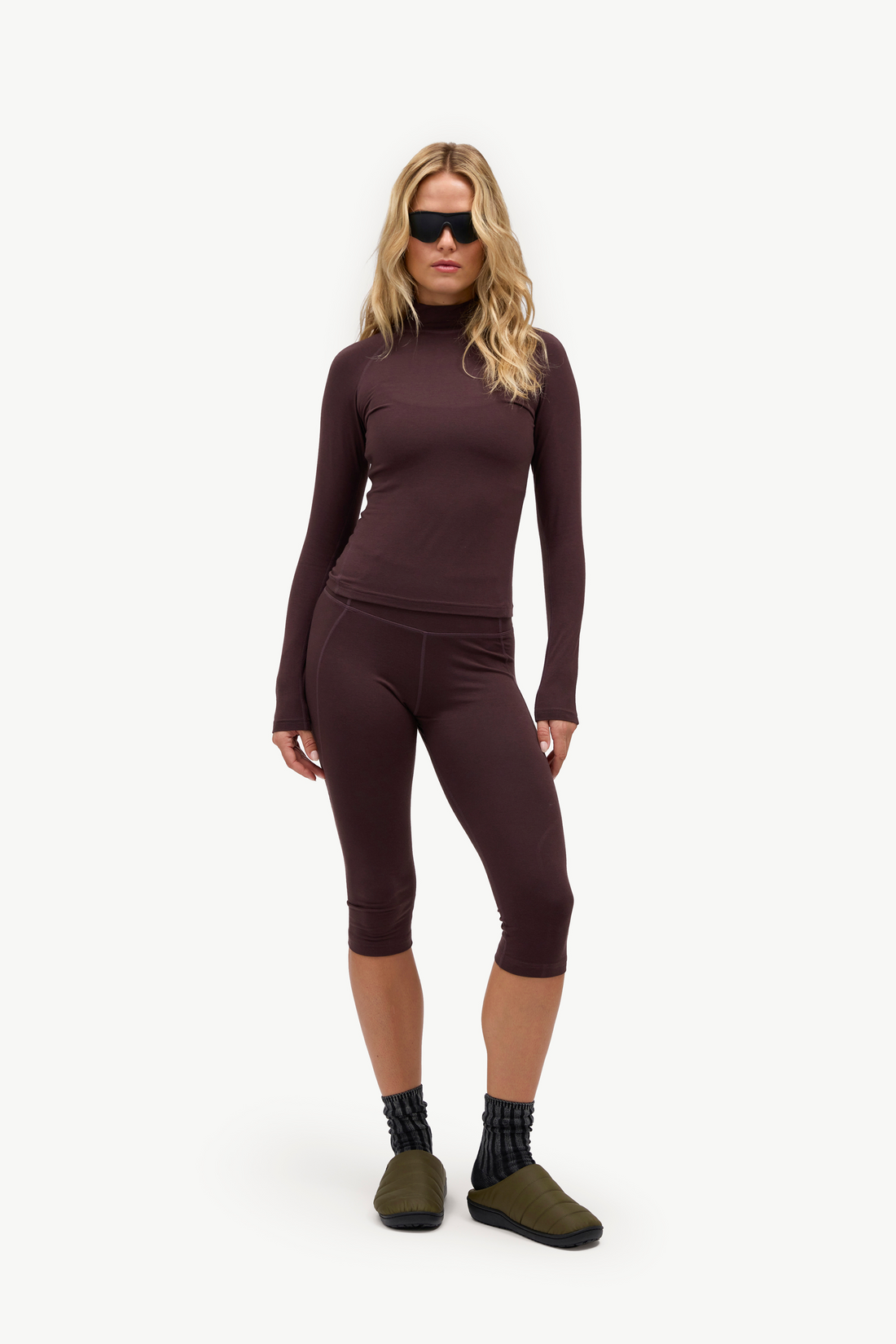 A woman modeling a matching dark plum-brown ribbed knit loungewear set consisting of a long-sleeve mock neck top and capri leggings, styled with black socks and olive green slip-on shoes against a plain white studio background.
