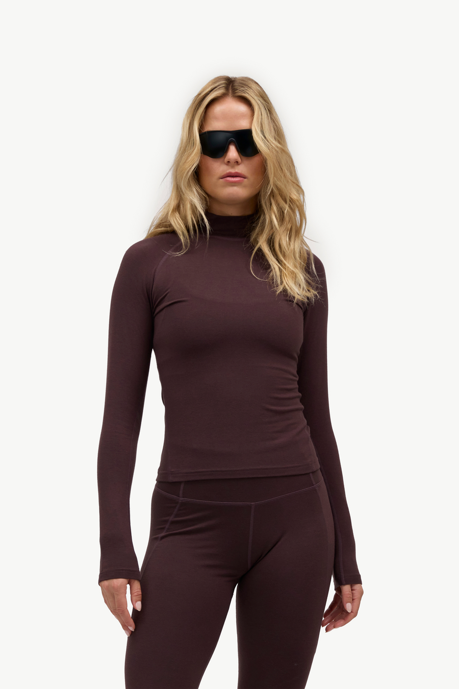 A woman modeling a dark plum long-sleeve mock neck top and matching leggings against a plain white studio background.