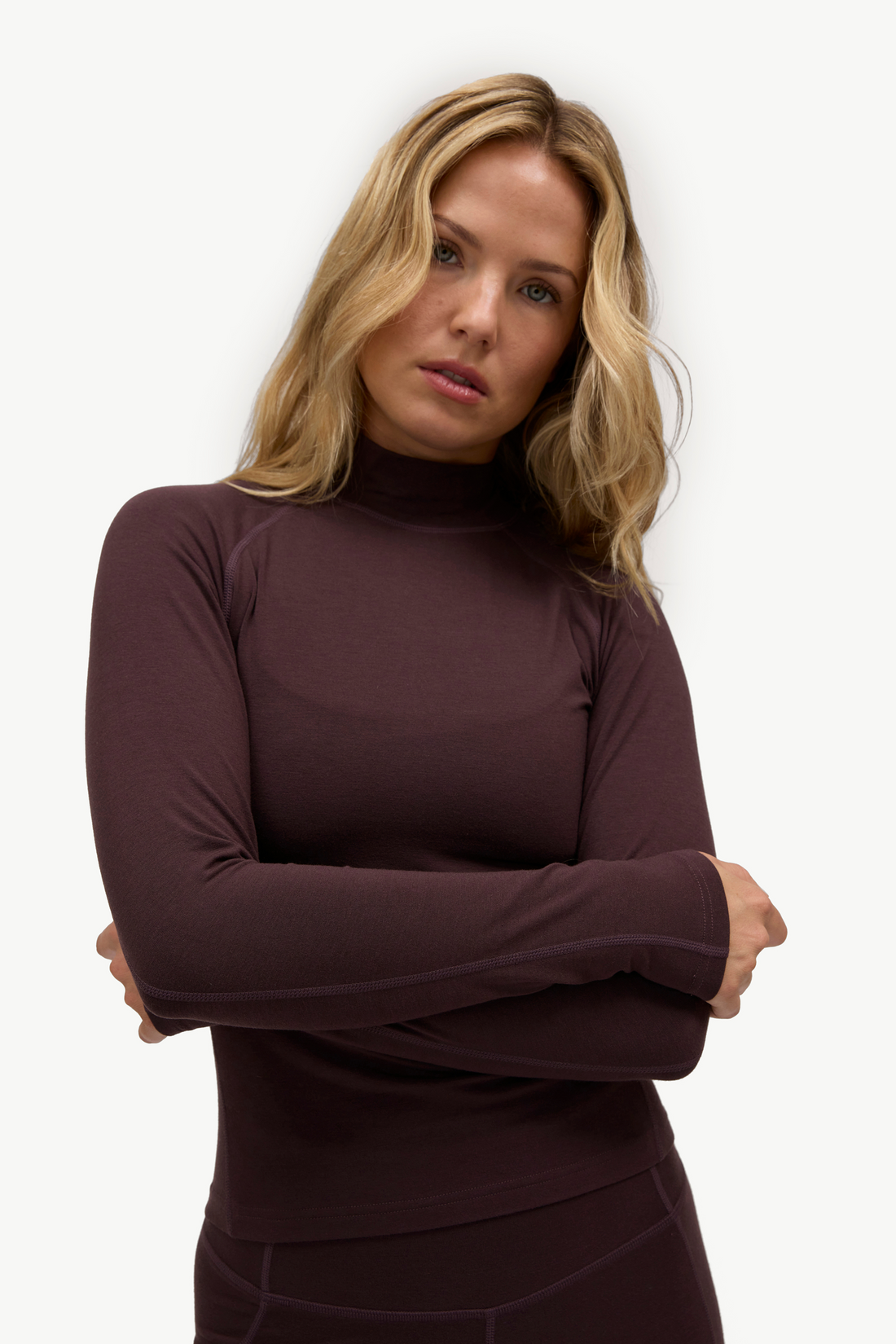 A blonde woman wearing a dark plum long-sleeve mock neck top, standing with her arms crossed against a plain white background.