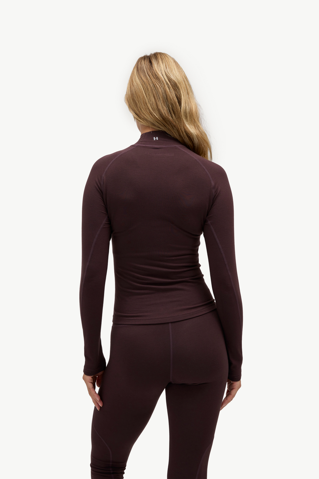 A woman shown from the back wearing a dark plum long-sleeve athletic top and matching leggings against a clean white studio background.