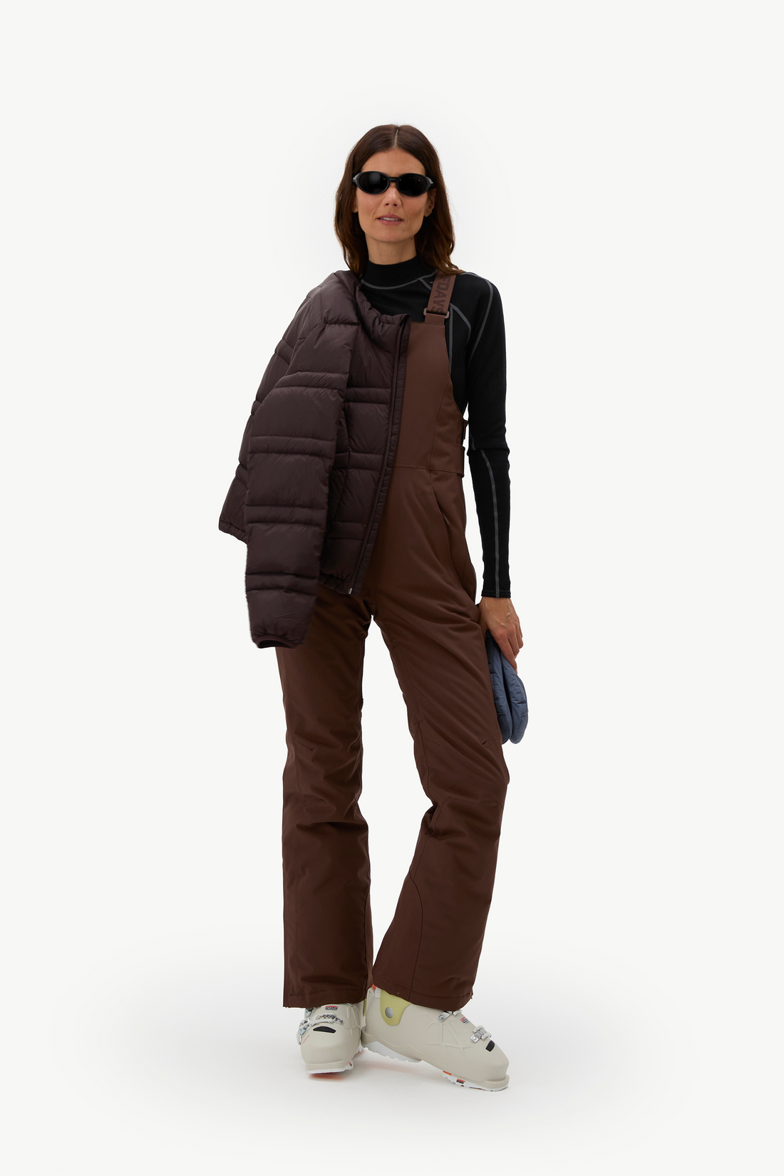 A model stands against a plain white studio background wearing a dark brown puffer vest over a black long-sleeve top and matching brown snow pants, with white ski boots and sunglasses.
