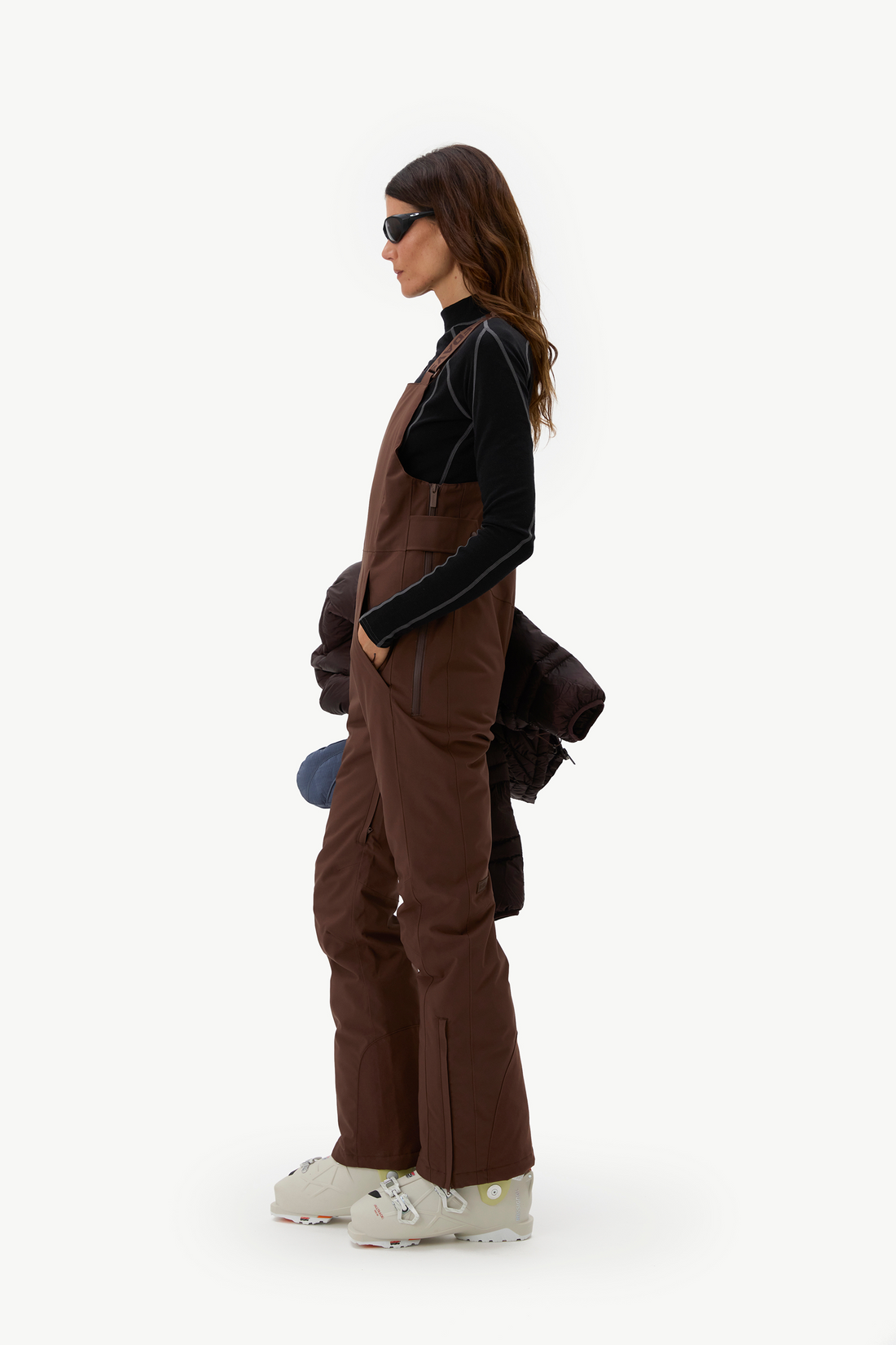 A woman stands in profile wearing brown snow bib overalls over a black long-sleeve top, with white snowboard boots, against a plain light gray studio background.