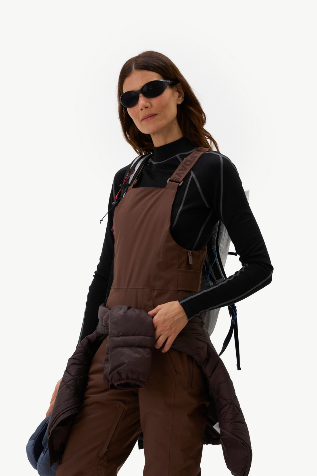 A woman modeling brown insulated outdoor overalls over a black long-sleeve top, wearing dark sunglasses and holding a dark puffer jacket, against a plain white background.