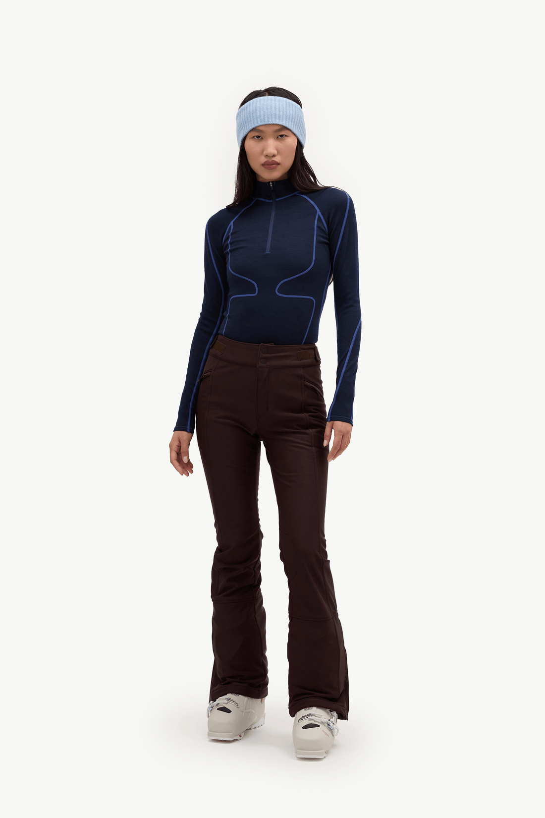A woman modeling a dark navy long-sleeve athletic top with light blue contour piping, paired with dark brown flared ski pants, a light blue headband, and white sneakers, set against a plain white studio background.