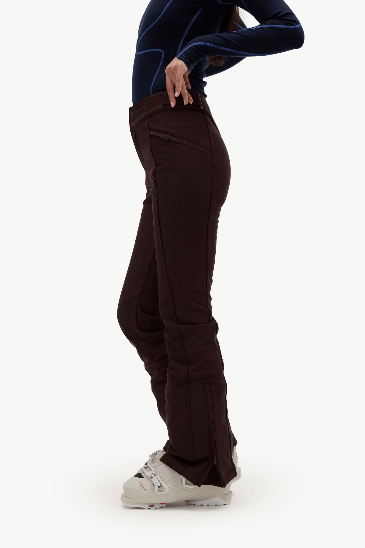 A side-profile view of a person wearing dark brown ski pants, a dark navy long-sleeve athletic top, and white ski boots against a plain white studio background.
