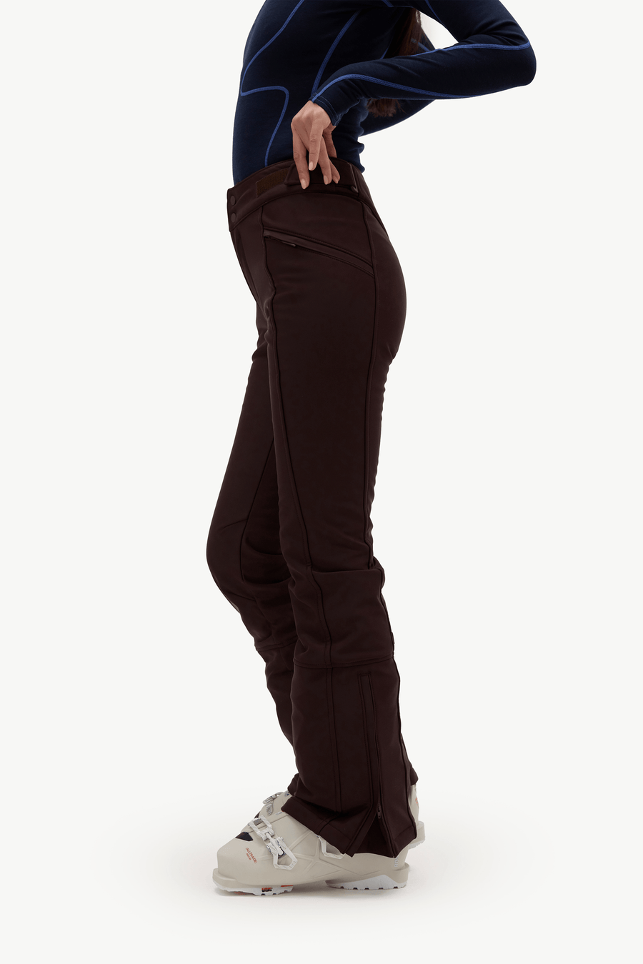 A side-profile view of a person wearing dark brown ski pants, a dark navy long-sleeve athletic top, and white ski boots against a plain white studio background.