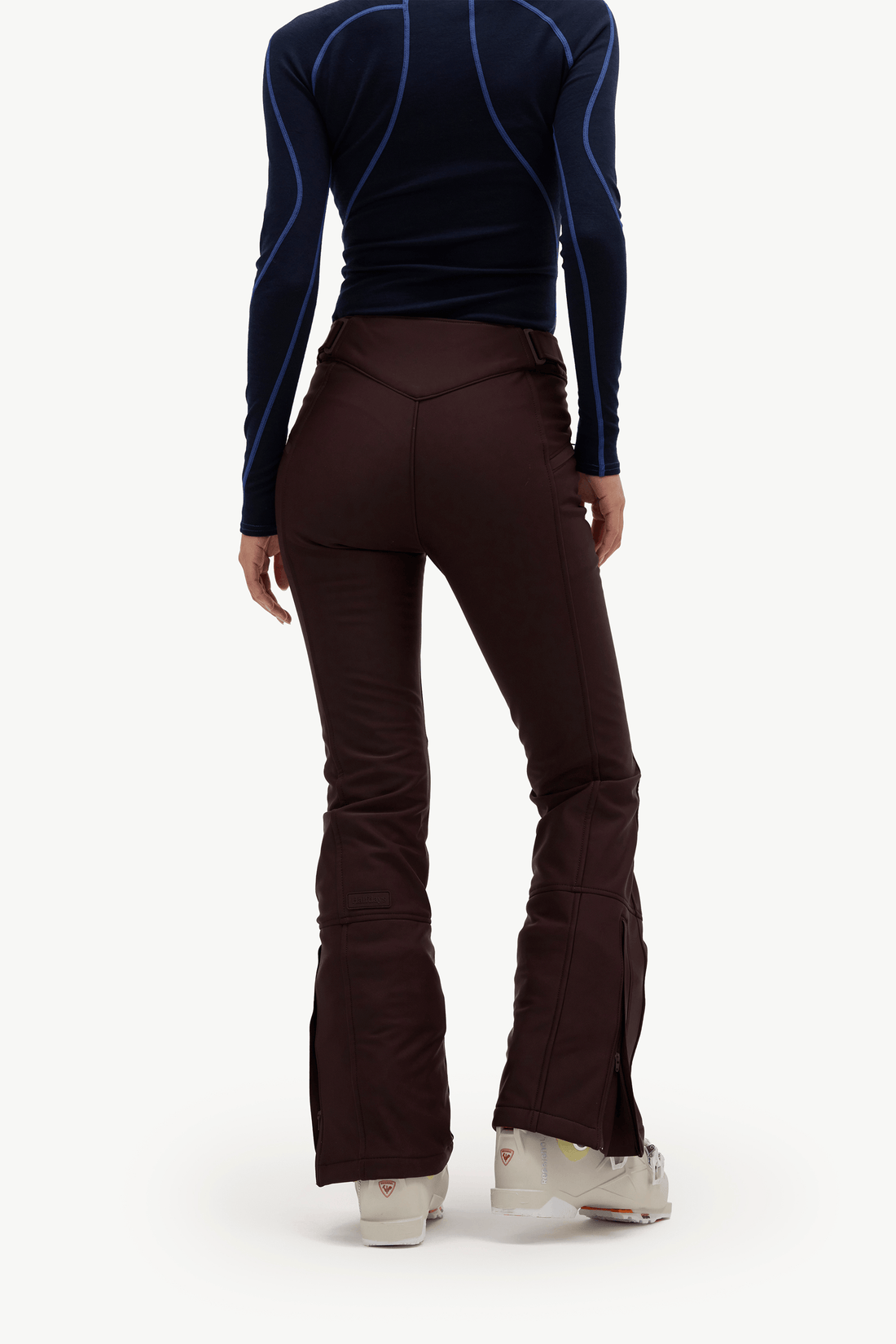 Rear view of a person wearing dark brown flared ski pants, a navy long-sleeve athletic top with blue piping, and white ski boots against a light gray studio background.