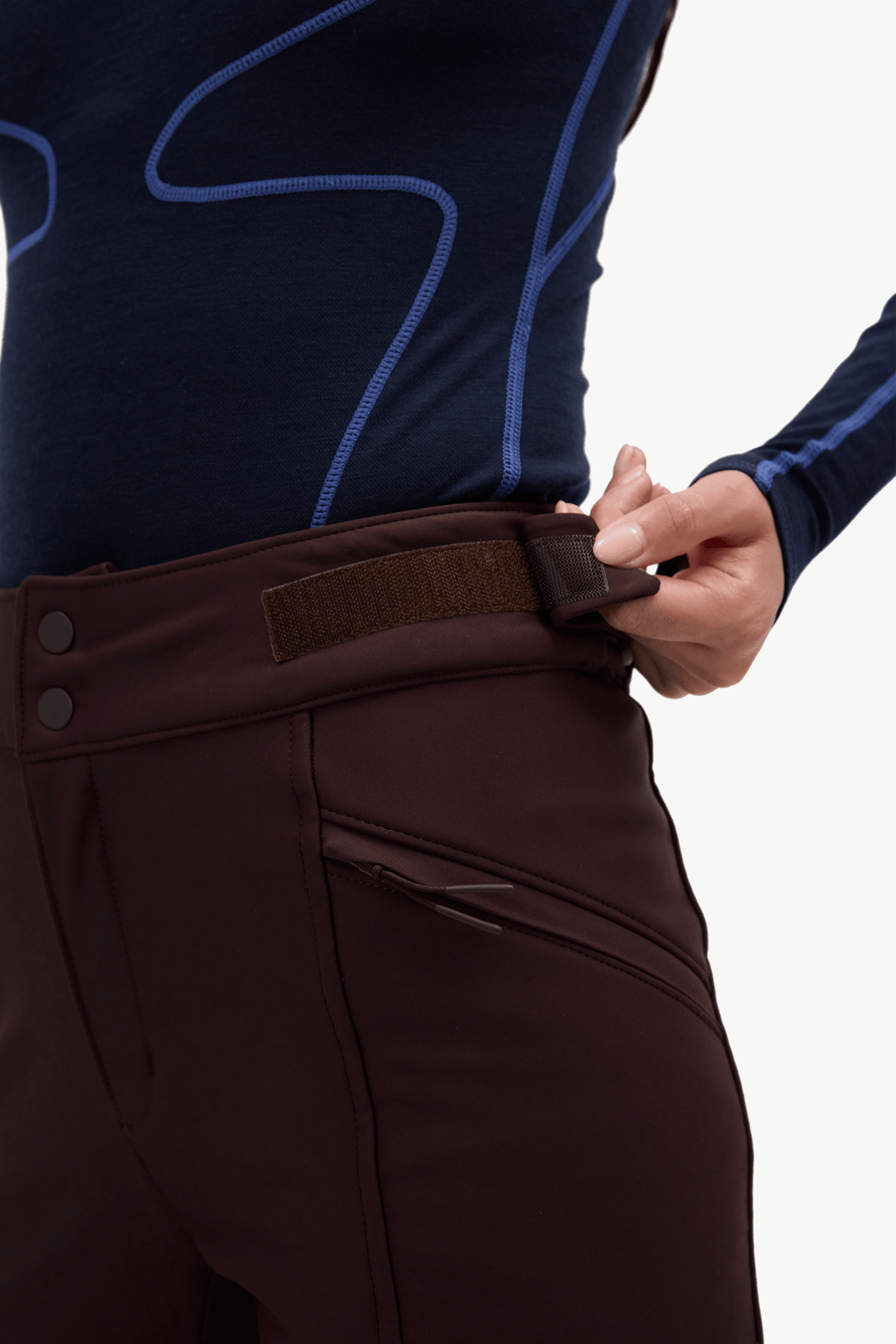 Close-up of a person wearing dark brown riding breeches and a navy long-sleeve riding shirt, adjusting the waistband of the breeches with one hand.