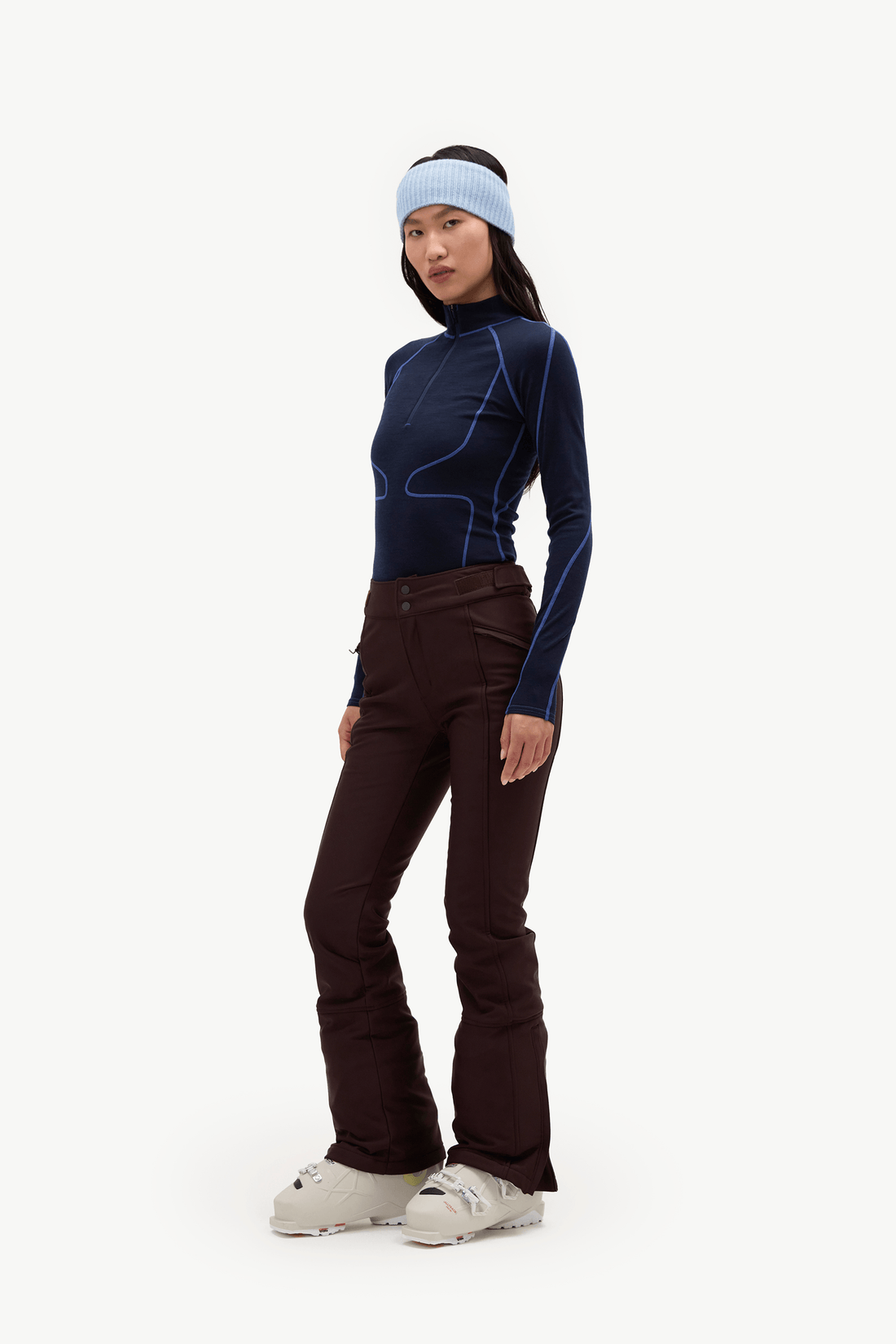 A model wearing a dark navy long-sleeve athletic top with bright blue contour lines, dark brown ski pants, a light blue headband, and white ski boots, standing against a solid white background.