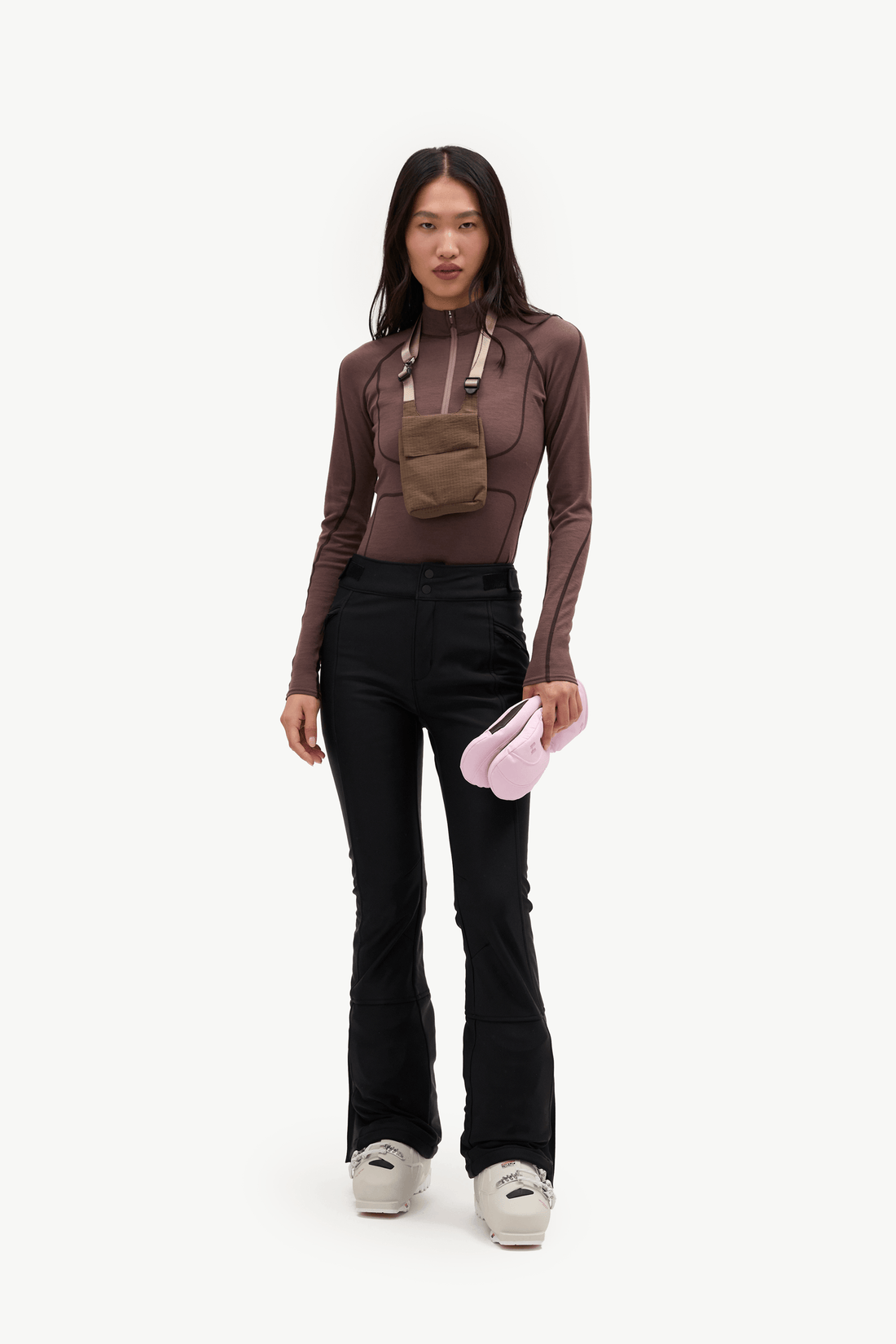 A model stands against a plain white studio background wearing a fitted brown mock-neck top with a small brown crossbody pouch, black flared pants, and light gray sneakers, while holding a pink item in one hand.