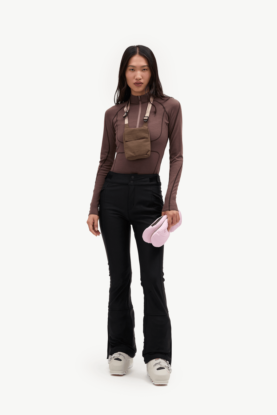 A model stands against a plain white studio background wearing a fitted brown mock-neck top with a small brown crossbody pouch, black flared pants, and light gray sneakers, while holding a pink item in one hand.