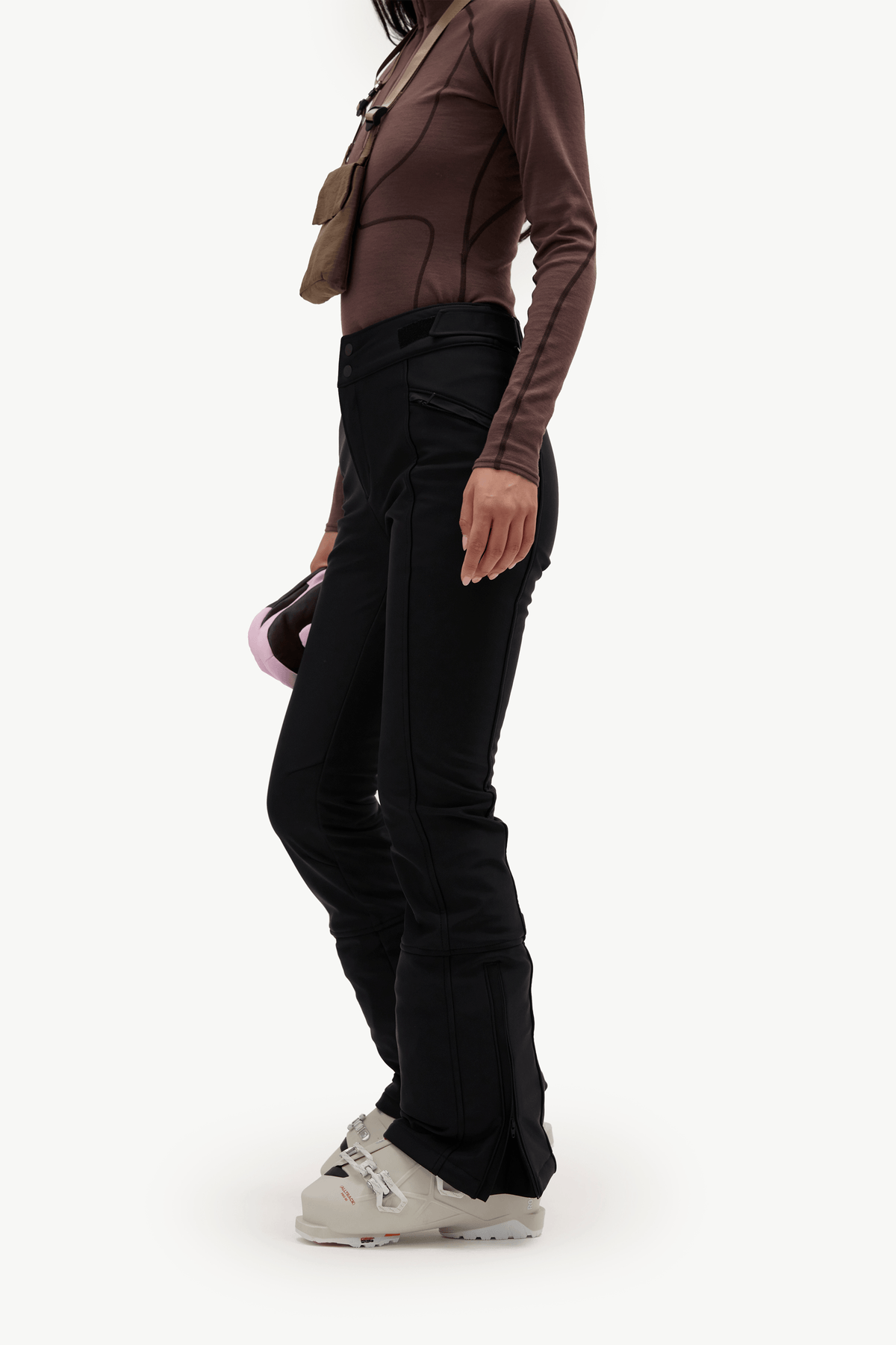 A side-profile view of a model wearing black ski pants, a brown long-sleeve top, and light beige ski boots against a plain white studio background.
