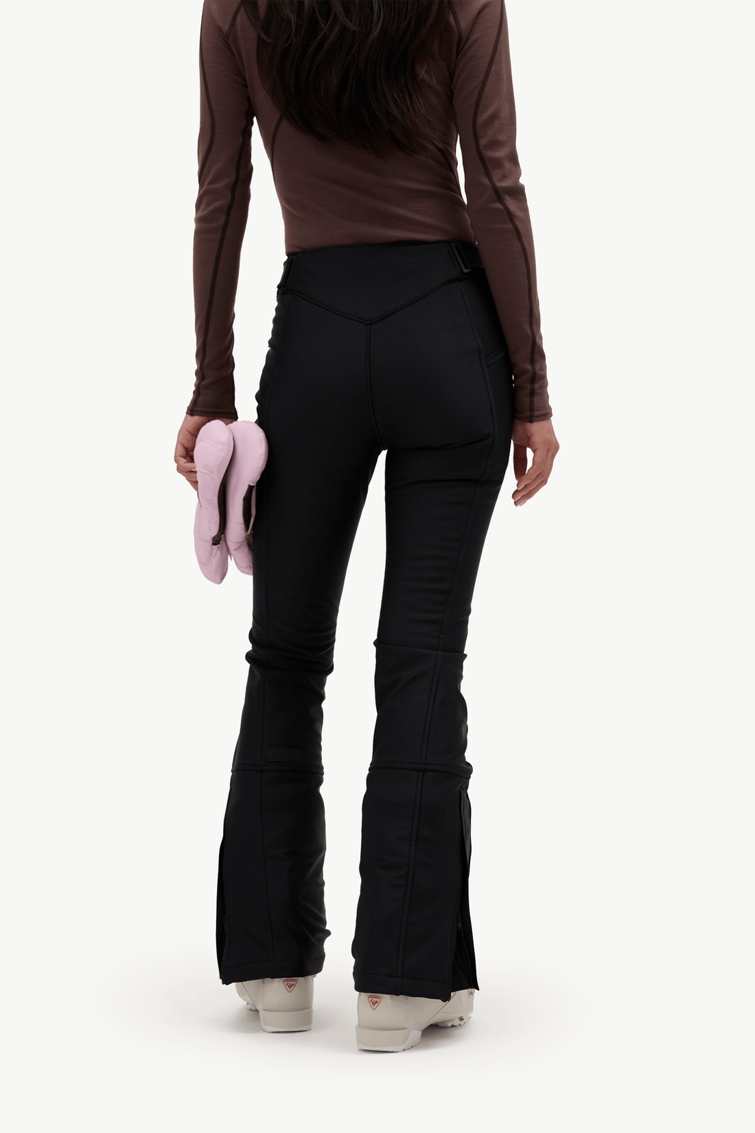 Rear view of a model wearing black flared pants with side zipper details, a brown long-sleeve top, and white sneakers, holding a pink item against a plain white background.
