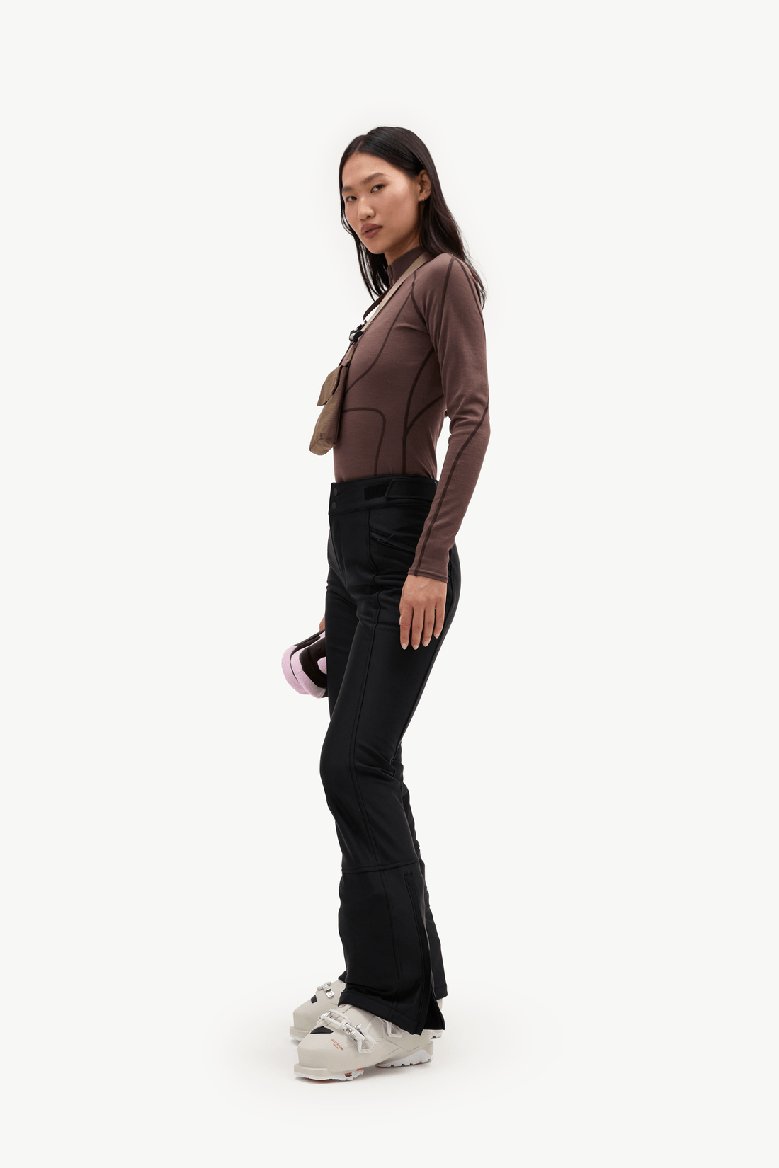 A woman stands in profile wearing a fitted brown long-sleeve top, black pants, and white sneakers against a plain light gray studio background.