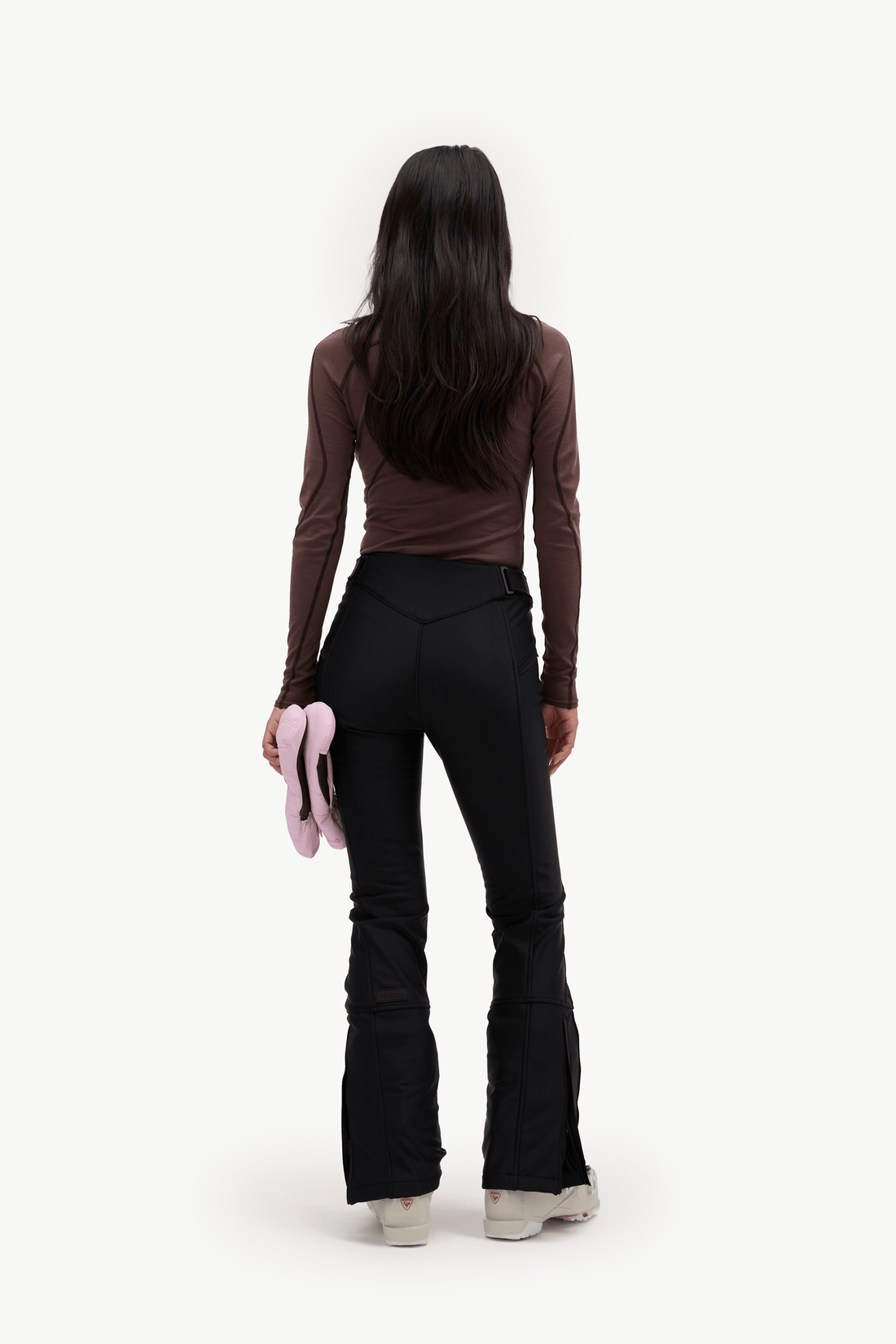A model is shown from behind wearing a brown long-sleeve top and black flared pants, holding a pink item in one hand against a plain white background.