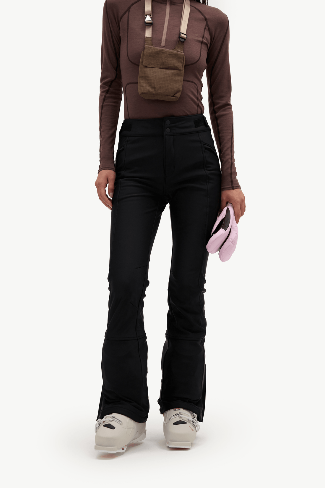 A model wearing black flared pants, a brown long-sleeve top, and a brown crossbody bag, holding a pink hat against a plain white background.