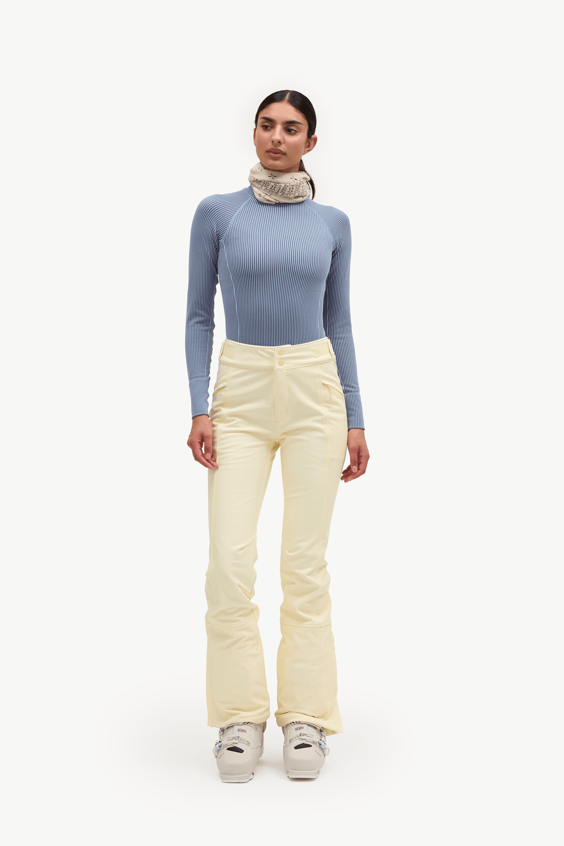 A model stands against a plain white studio background wearing a light blue long-sleeve top, cream-colored ski pants, and white sneakers.