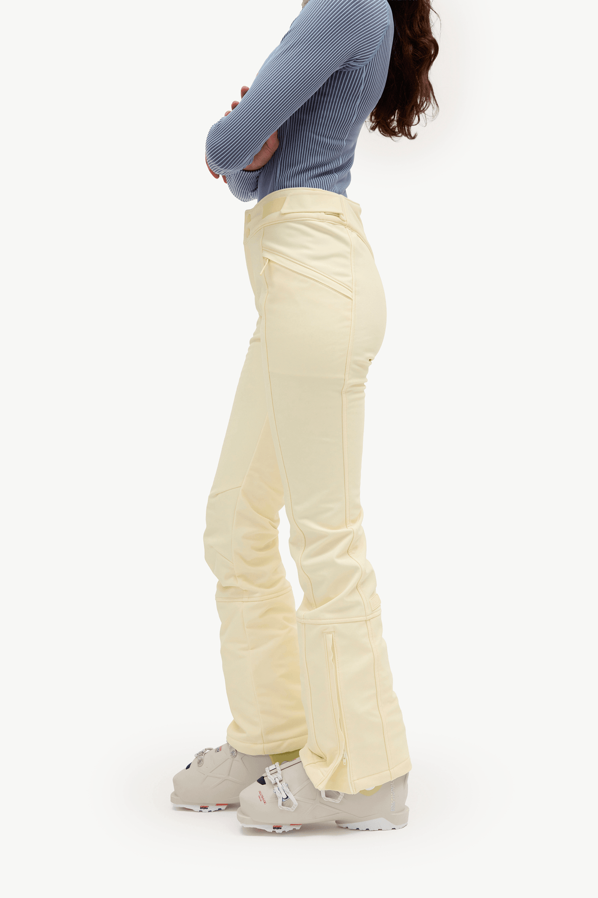 A side-profile view of a model wearing light cream flared ski pants, a blue ribbed long-sleeve top, and ski boots against a plain white studio background.