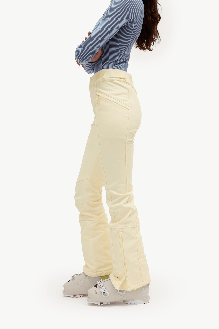 A side-profile view of a model wearing light cream flared ski pants, a blue ribbed long-sleeve top, and ski boots against a plain white studio background.