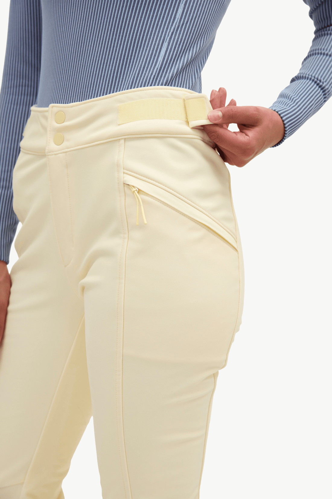 Close-up of a person wearing cream-colored riding breeches with a zippered pocket and button waist, paired with a blue striped shirt against a white background.