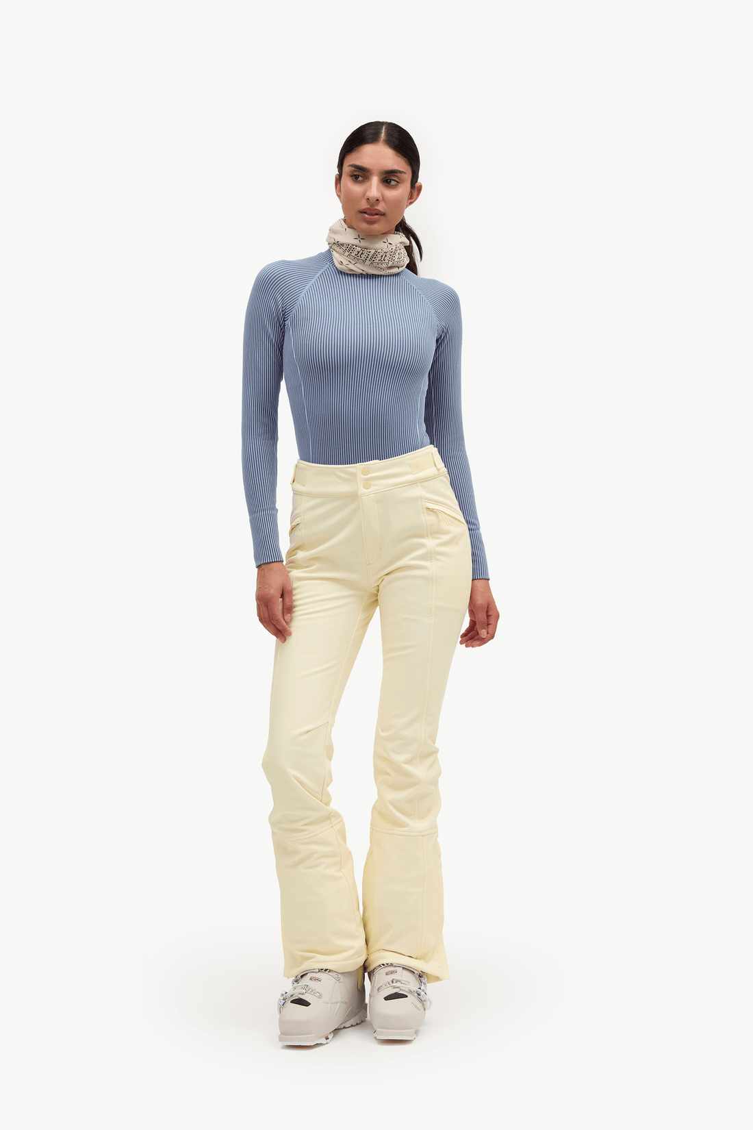 A model stands against a plain white studio background wearing a light blue long-sleeve mock-neck top, cream-colored ski pants, and white ski boots.