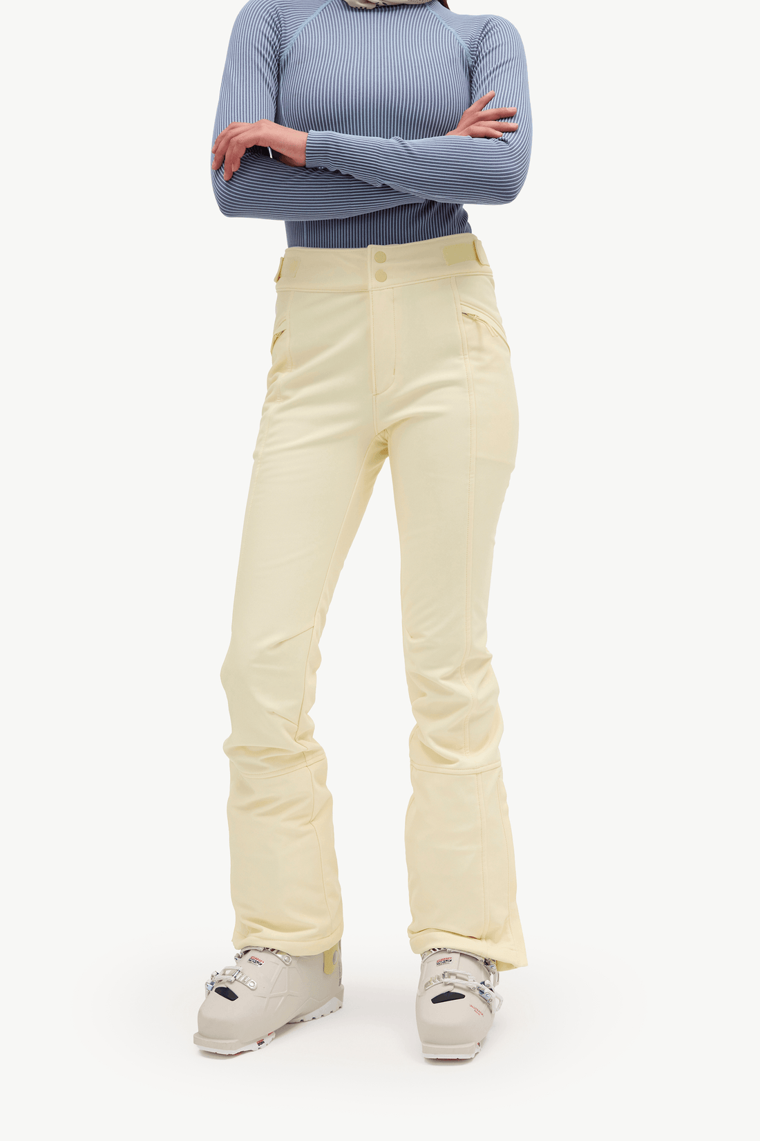 A model wearing light yellow ski pants, a dusty blue ribbed long-sleeve top, and off-white snowboard boots, standing with arms crossed against a plain white studio background.
