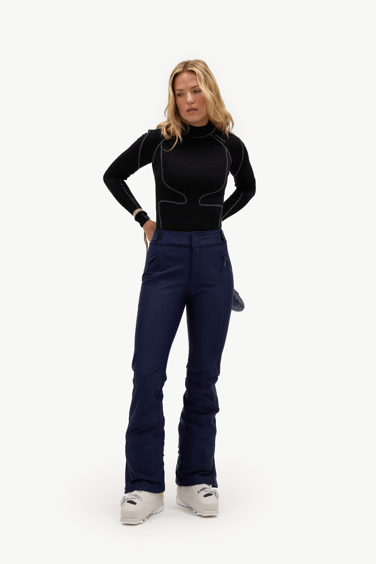 A woman modeling a black long-sleeve athletic top and navy blue ski pants with white ski boots, standing against a plain white studio background.