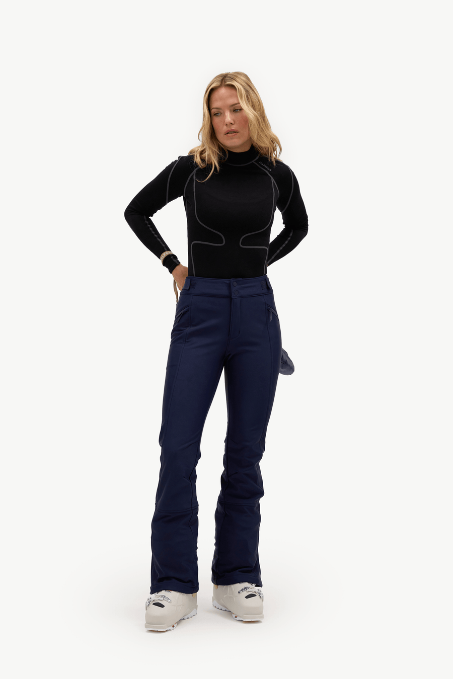 A woman modeling a black long-sleeve athletic top and navy blue ski pants with white ski boots, standing against a plain white studio background.