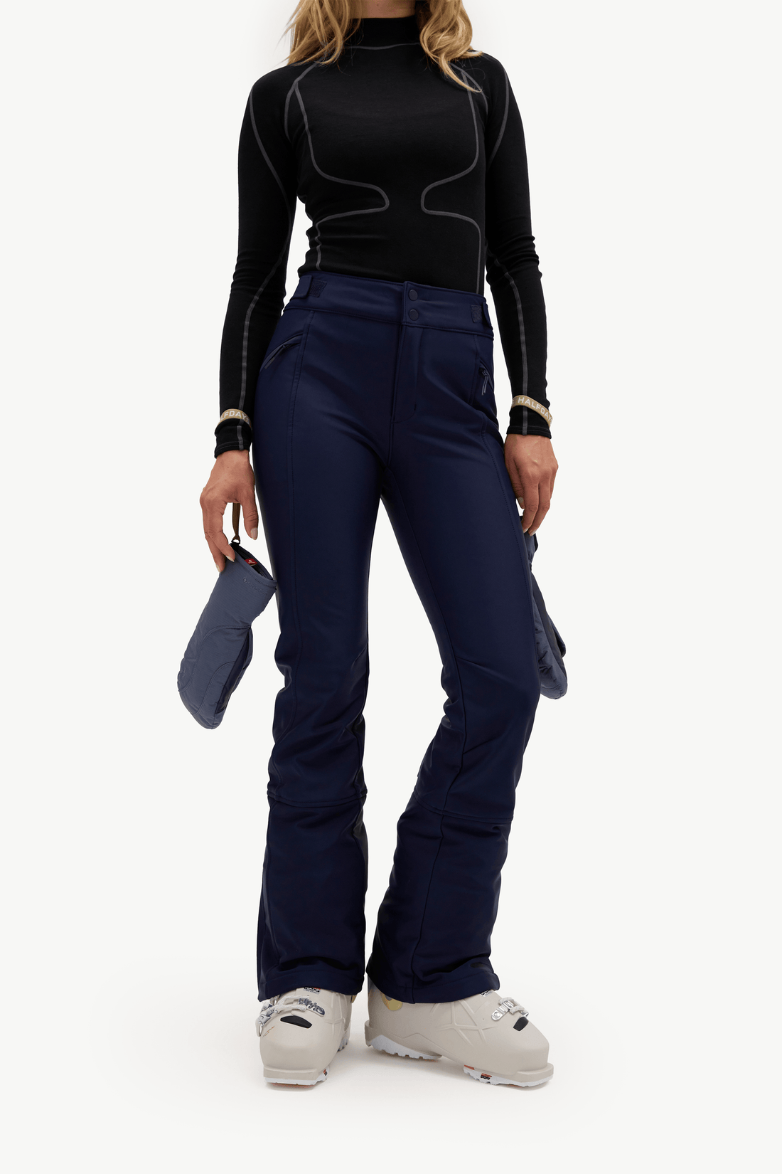 A model wearing dark navy ski pants, a black long-sleeve top, and white ski boots, holding a pair of dark blue gloves against a plain white studio background.