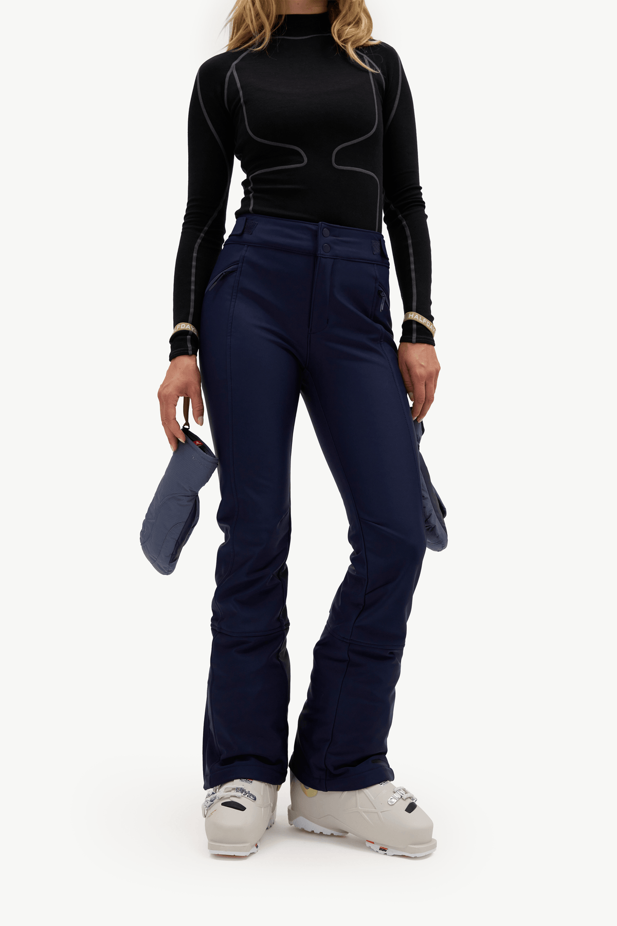 A model wearing dark navy ski pants, a black long-sleeve top, and white ski boots, holding a pair of dark blue gloves against a plain white studio background.
