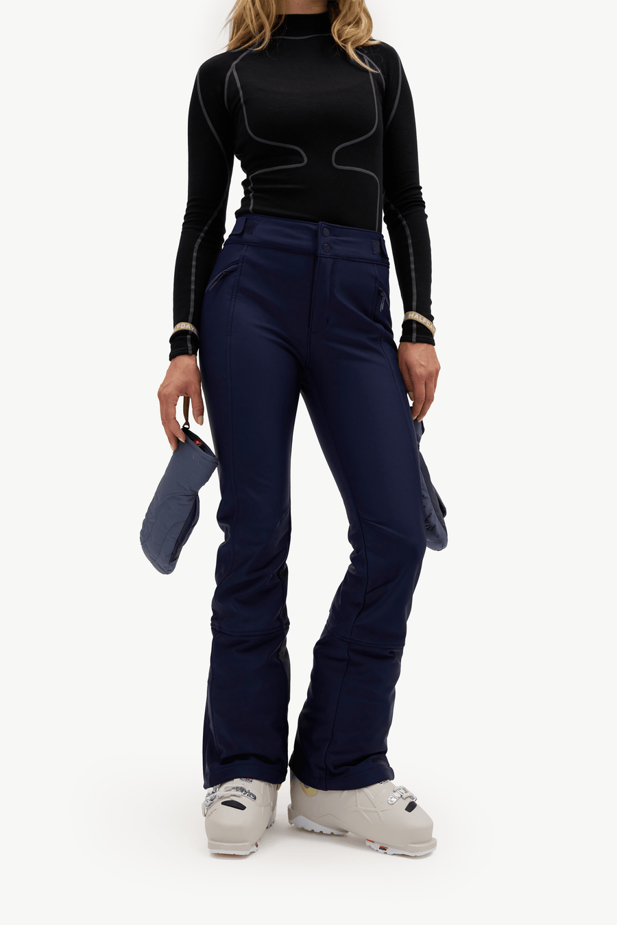 A model wearing dark navy ski pants, a black long-sleeve top, and white ski boots, holding a pair of dark blue gloves against a plain white studio background.