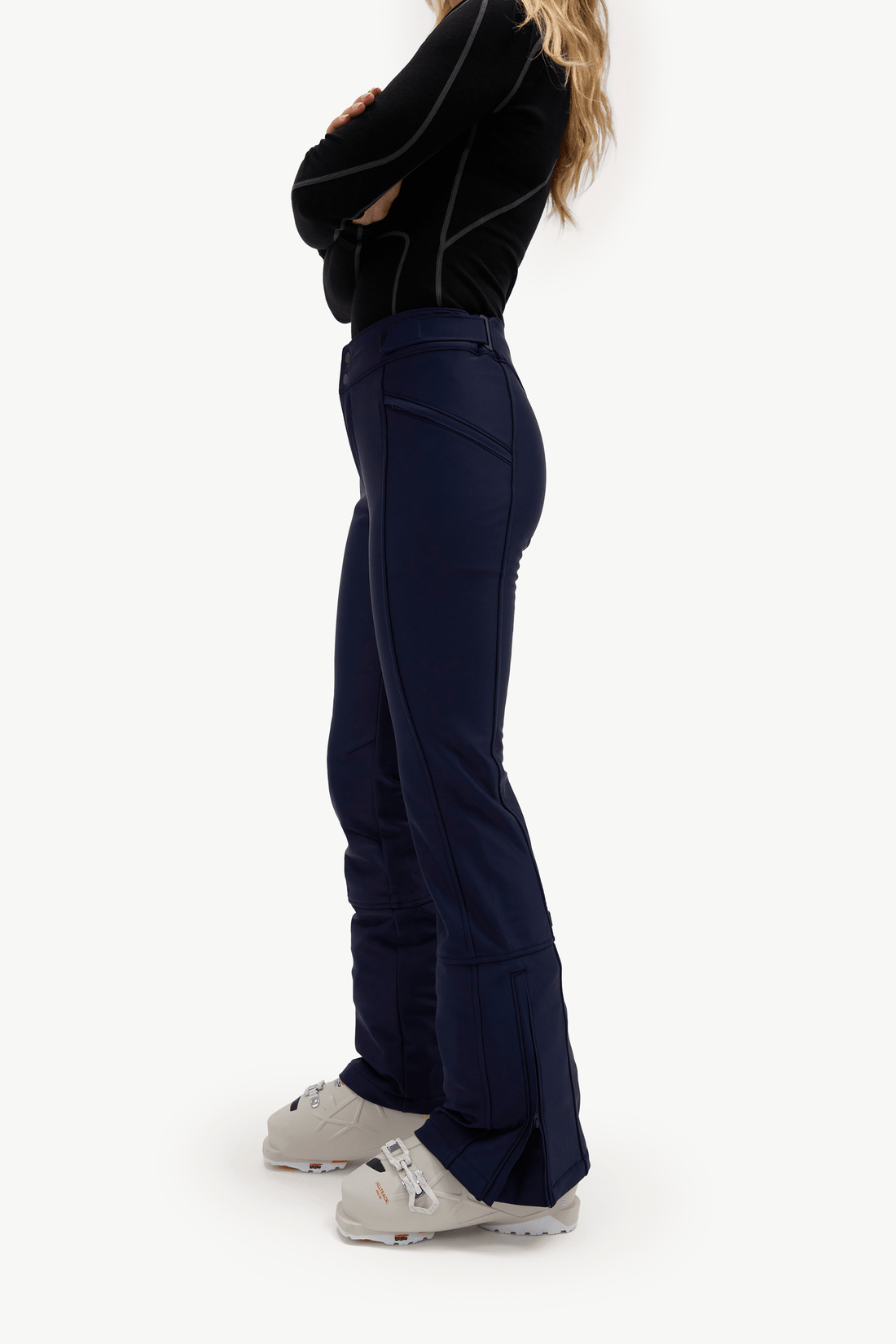 A side-profile view of a model wearing dark navy ski pants with black piping details, a black long-sleeve top, and light gray ski boots, set against a plain white studio background.