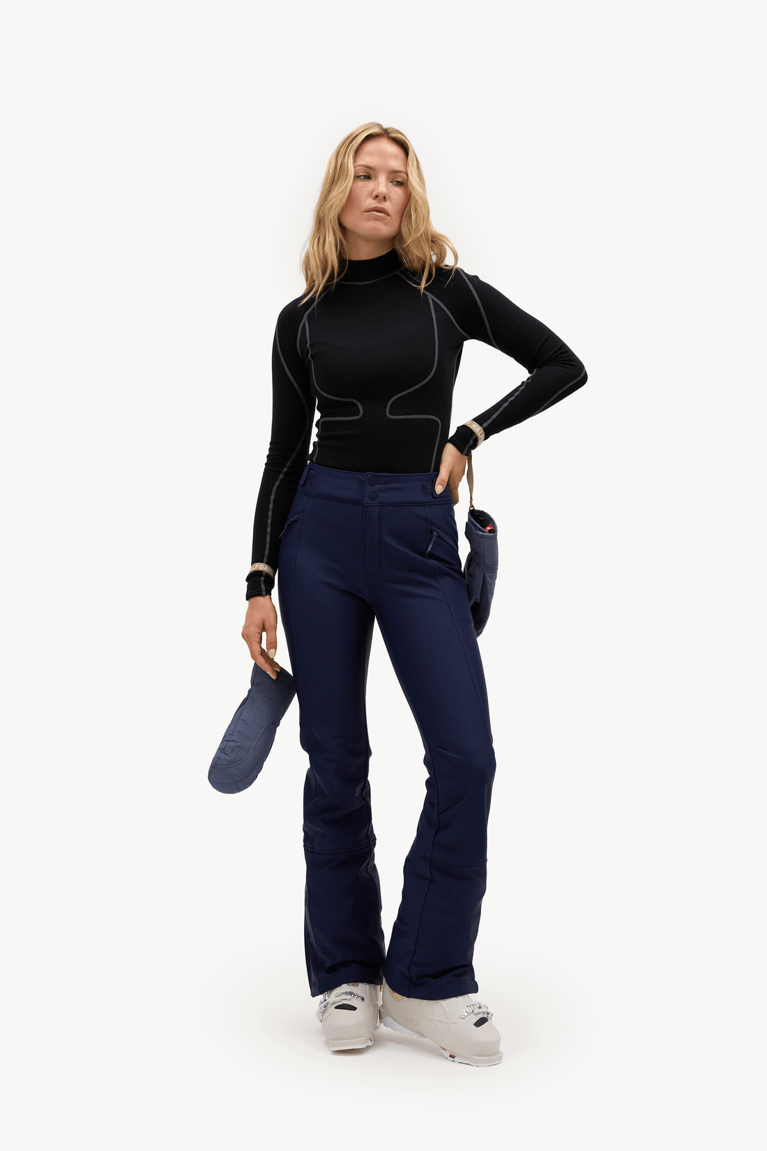 A woman modeling a black long-sleeve athletic top and navy blue flared ski pants, holding a navy blue ski boot against a clean white studio background.