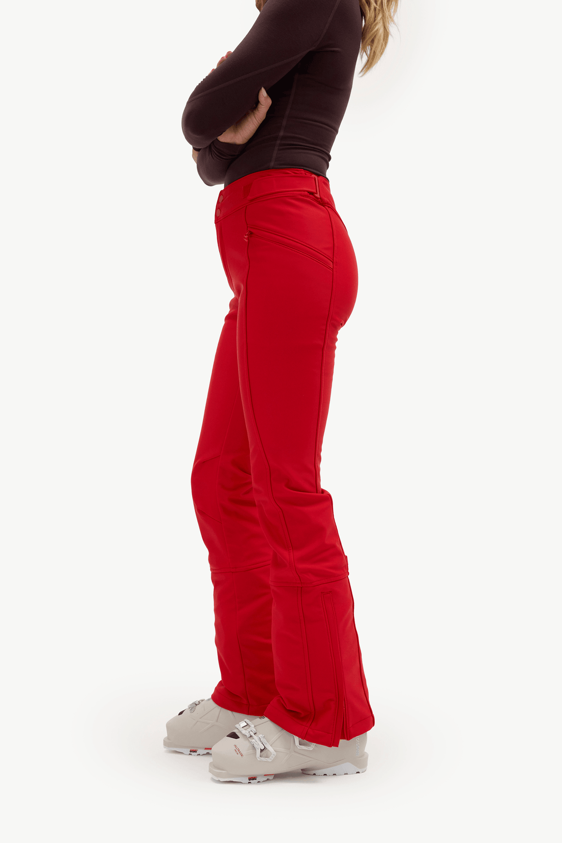 A side-profile view of a model wearing bright red ski pants, a dark brown long-sleeve top, and light gray ski boots against a plain white studio background.