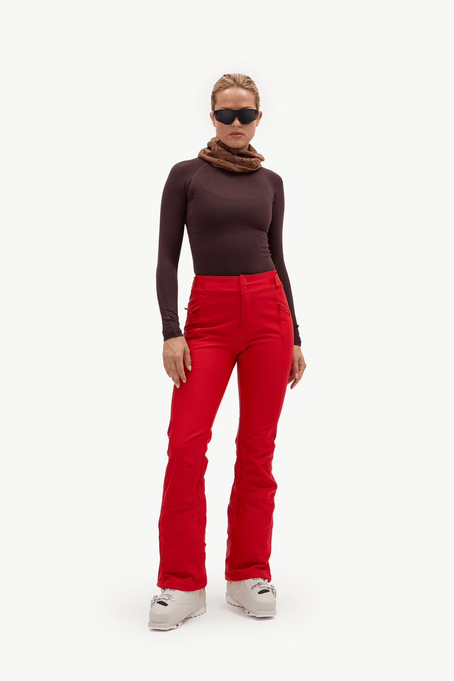 A model wearing bright red ski pants, a dark brown long-sleeve top, a brown knit neck warmer, and black sunglasses, standing against a solid white background.