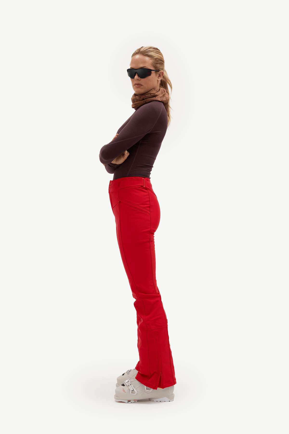 A woman stands in profile against a plain white studio background, wearing dark sunglasses, a dark plum long-sleeve top, bright red flared pants, a brown scarf, and white Croc sneakers.