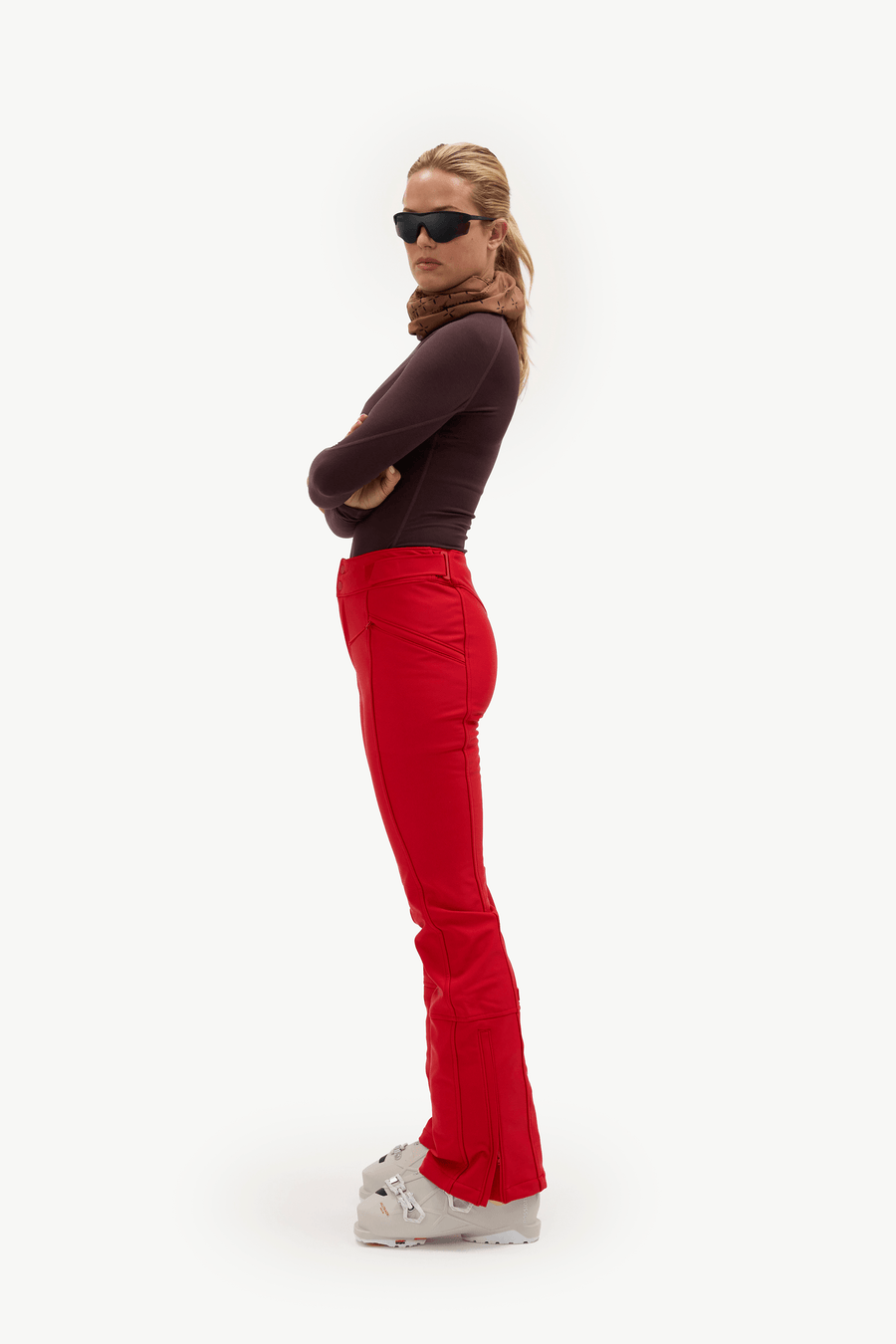 A woman stands in profile against a plain white studio background, wearing dark sunglasses, a dark plum long-sleeve top, bright red flared pants, a brown scarf, and white Croc sneakers.