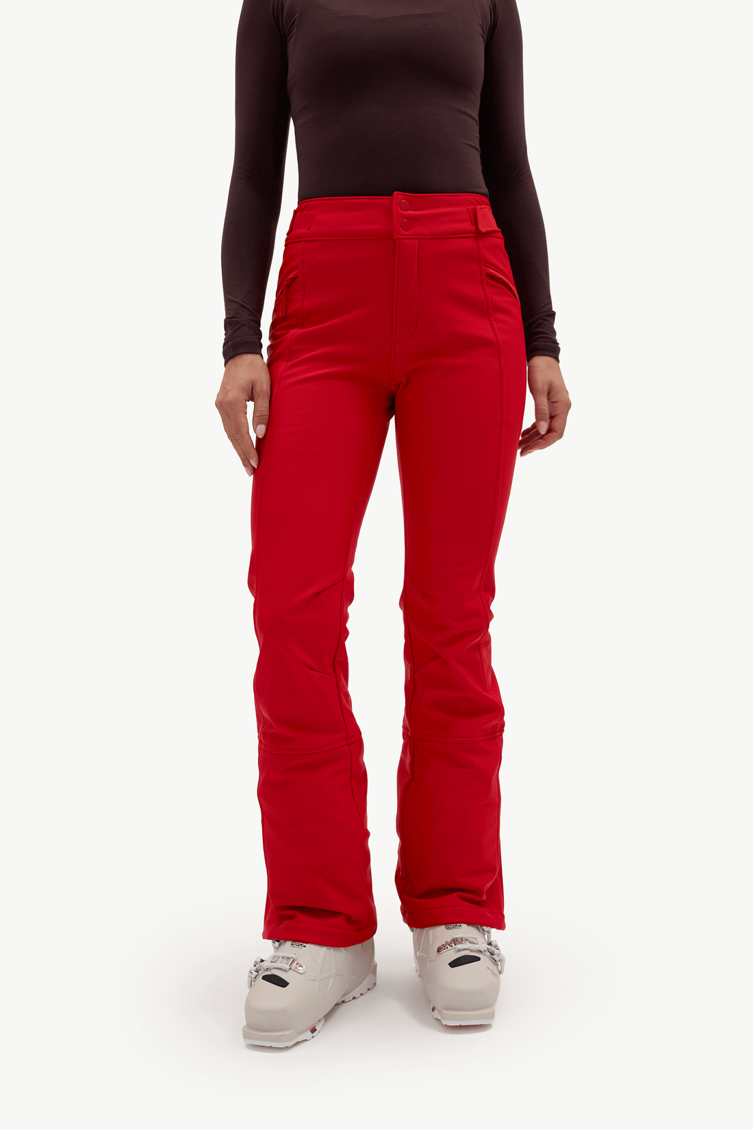 A model wearing bright red ski pants, a dark brown long-sleeve top, and light gray ski boots, standing against a plain white studio background.
