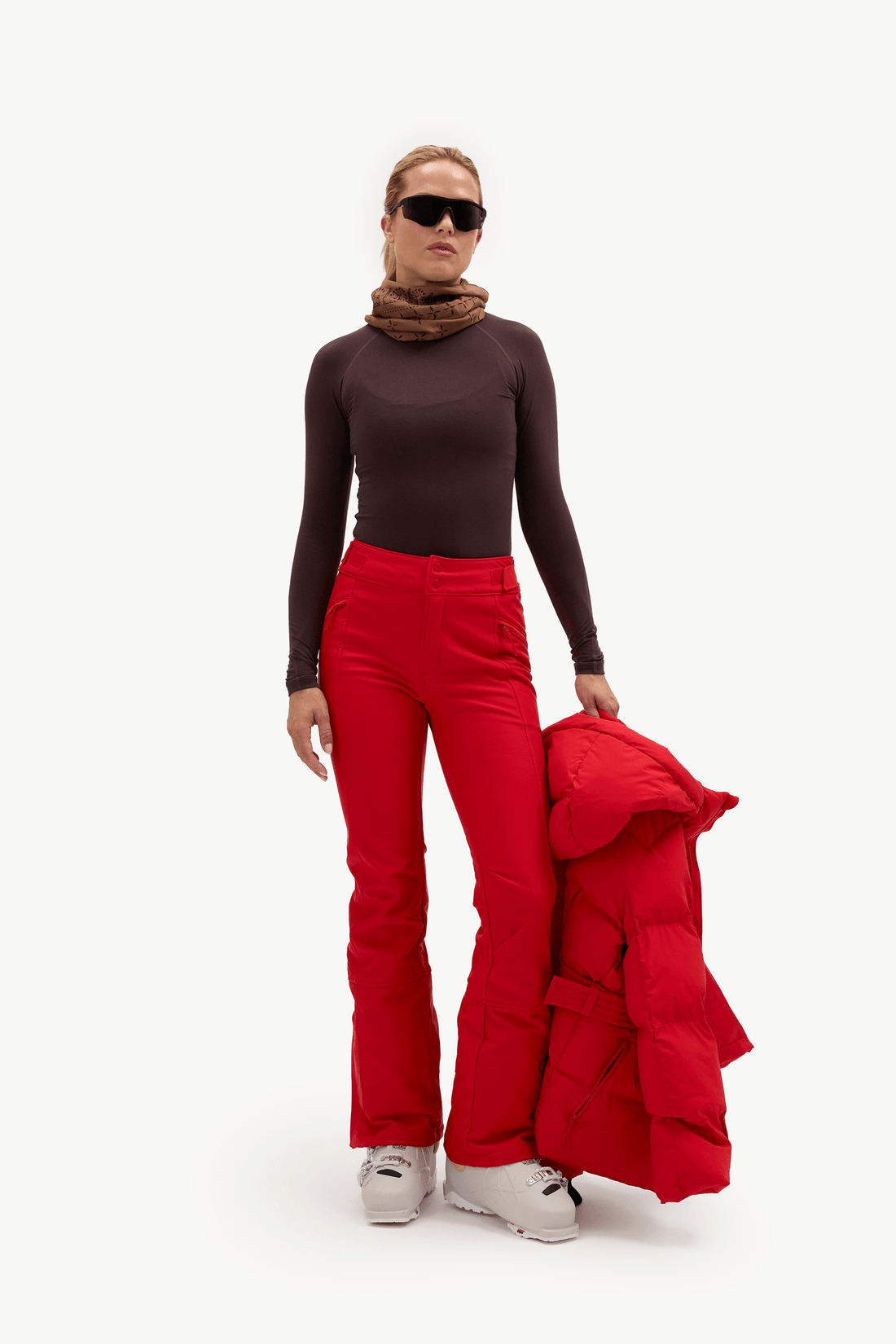 A model stands against a plain white studio background wearing bright red ski pants and a dark brown long-sleeve top, with a brown neck warmer, black sunglasses, and white ski boots. She is holding a folded red puffer jacket.