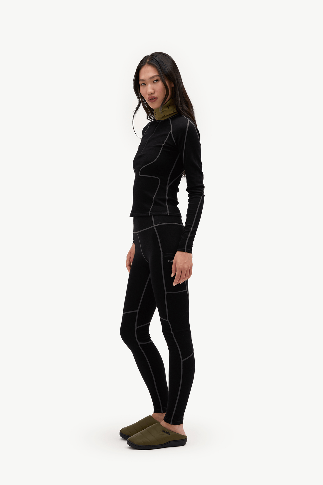 A woman modeling a black long-sleeve athletic top and matching black leggings with contrasting white seam lines, paired with olive green slip-on shoes, against a plain white studio background.