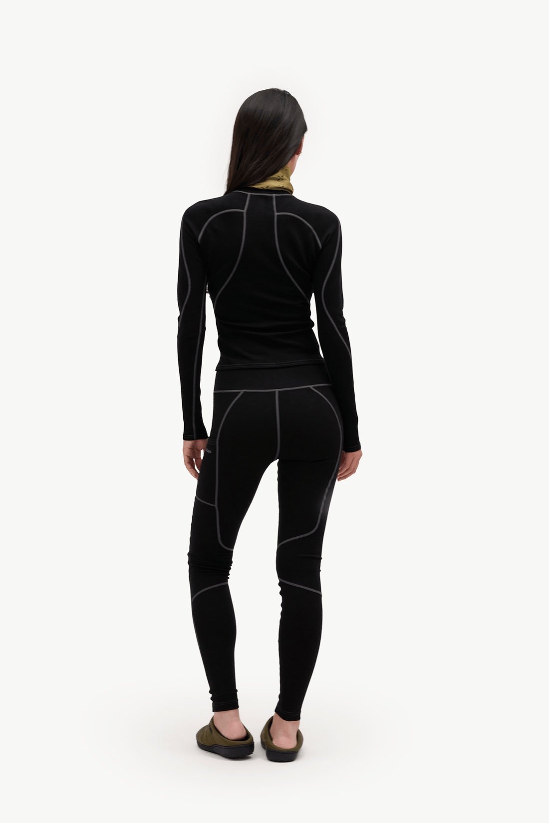 A woman viewed from behind wearing a black long-sleeve athletic top and matching black leggings with contrasting light gray seam lines, standing against a plain white studio background.