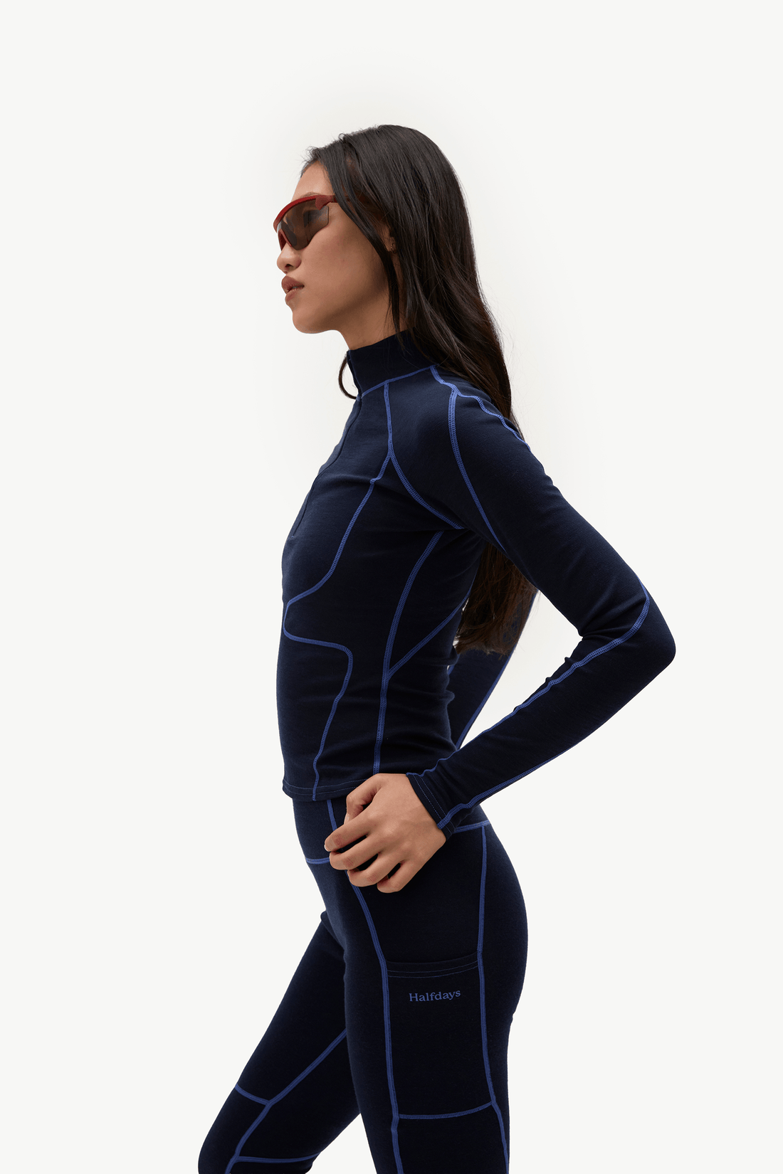 A woman in profile wearing a dark navy blue full-body wetsuit with bright blue seam detailing and matching dark leggings, photographed against a clean white studio background.