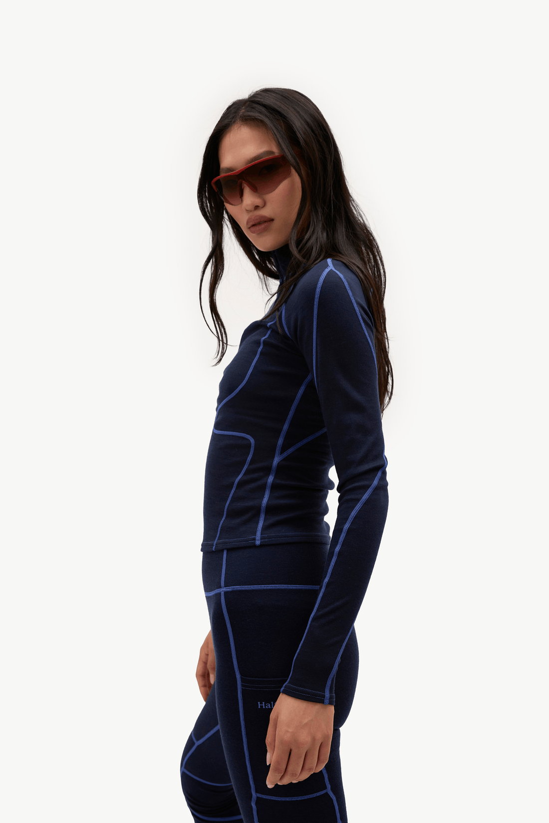 A woman in profile wearing a dark navy long-sleeve athletic top and matching leggings with bright blue contrast stitching, set against a plain white studio background.