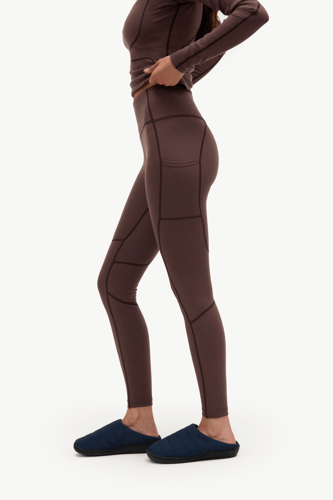 A model wearing dark brown high-waisted leggings with geometric paneling and a matching long-sleeve top, paired with navy blue slip-on slippers, shown from the waist down against a plain white background.